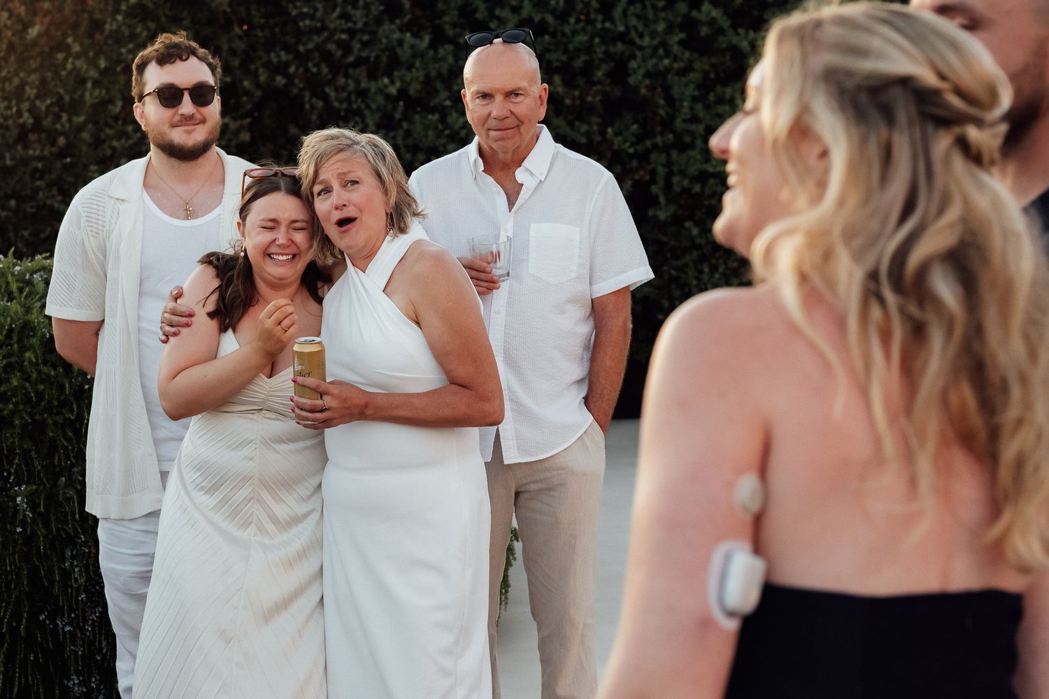 Group of guests in white attire smiling and laughing at an outdoor wedding celebration.