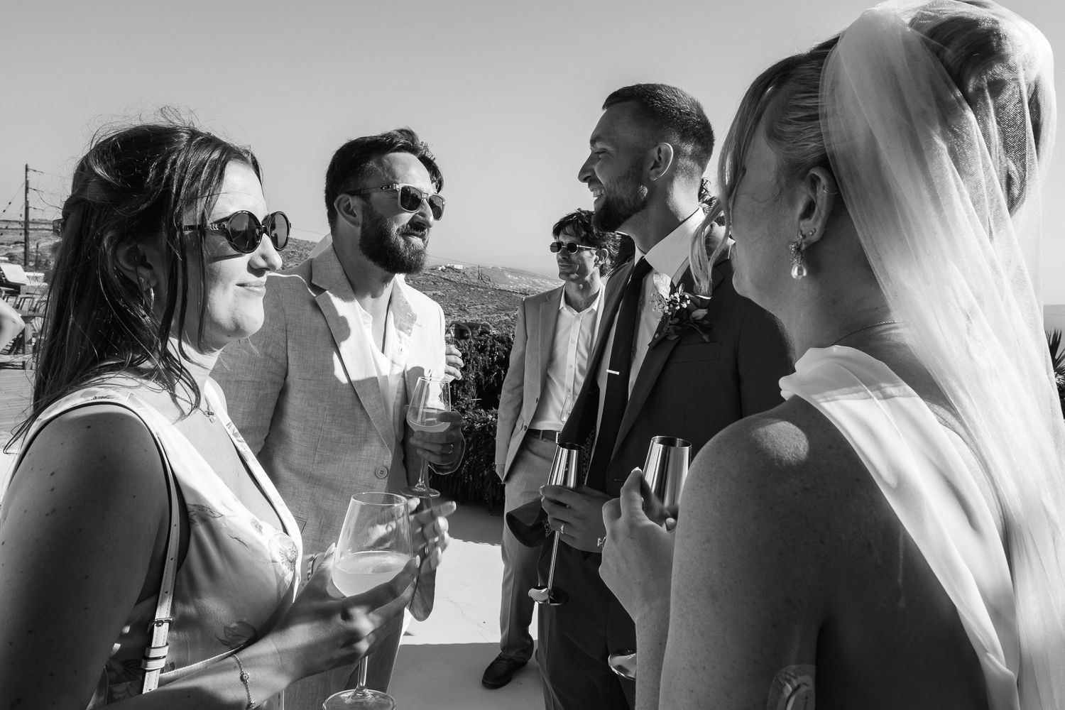 Black and white photo of elegantly dressed wedding guests socializing and laughing outdoors at a sunny reception.