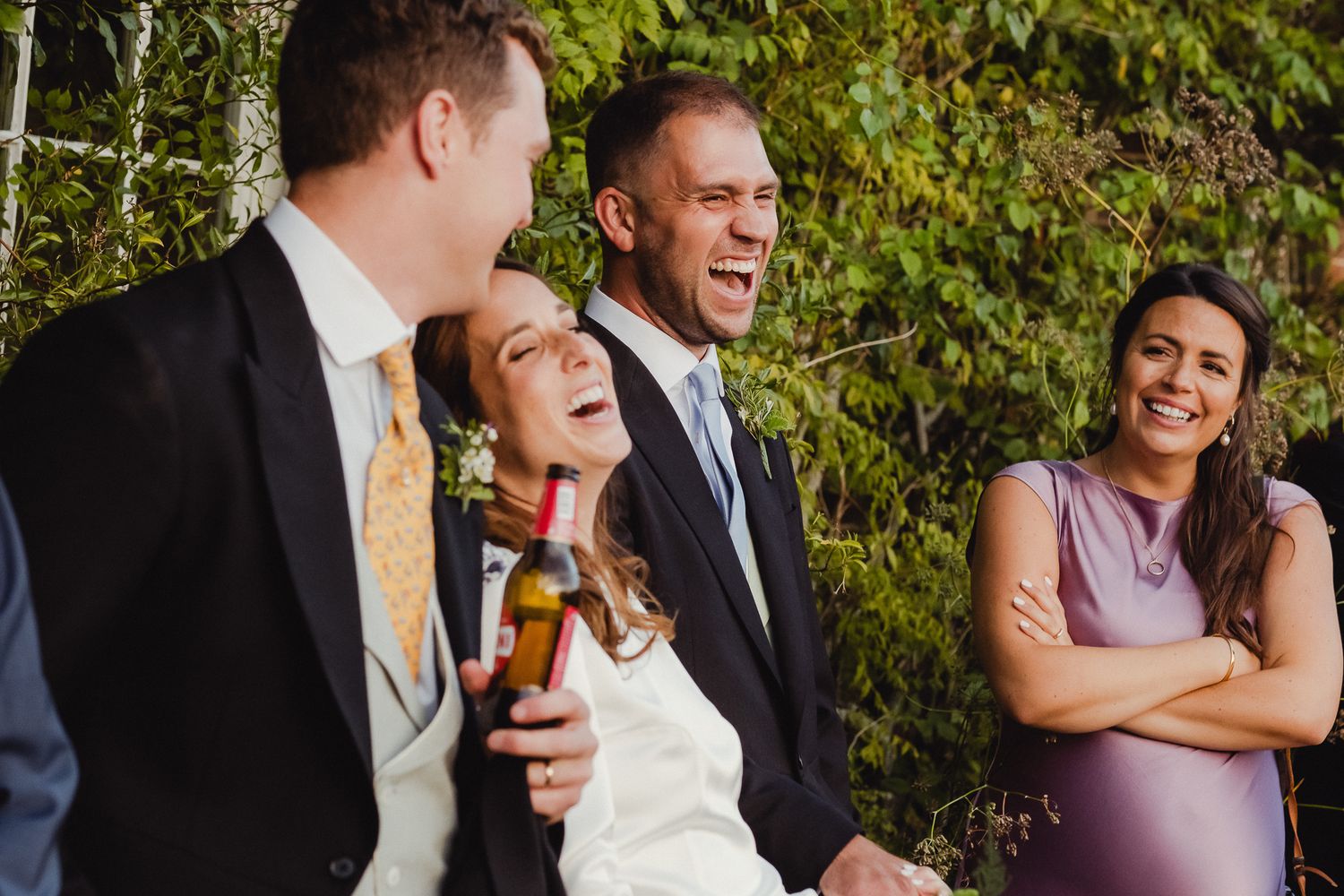 Wedding party laughs together while standing in a line outdoors with green foliage in the background.