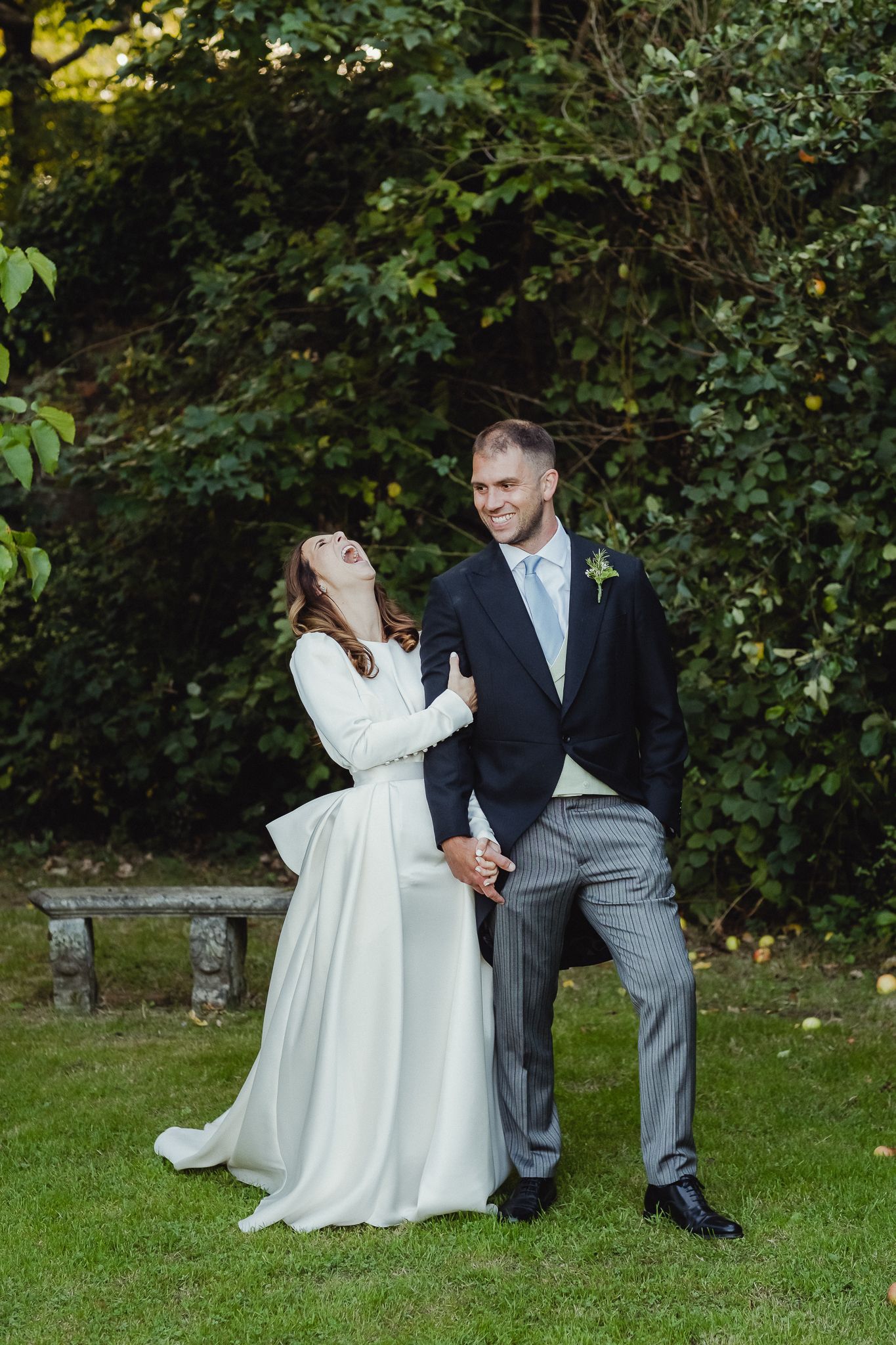 A couple in wedding attire playfully poses on grass with trees in the background during an outdoor photoshoot.