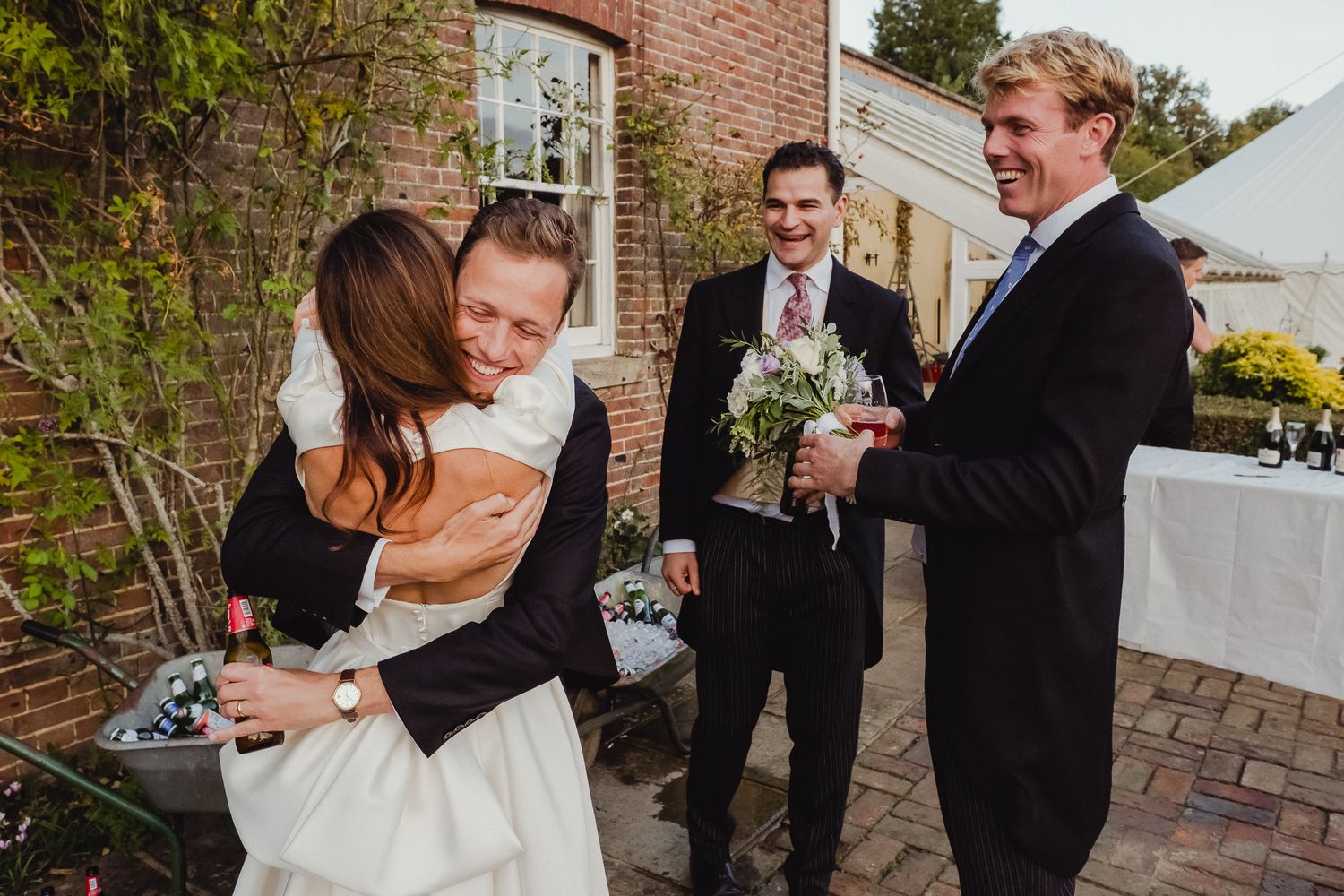 A joyful wedding celebration with people dancing and embracing on a brick patio during an outdoor reception.