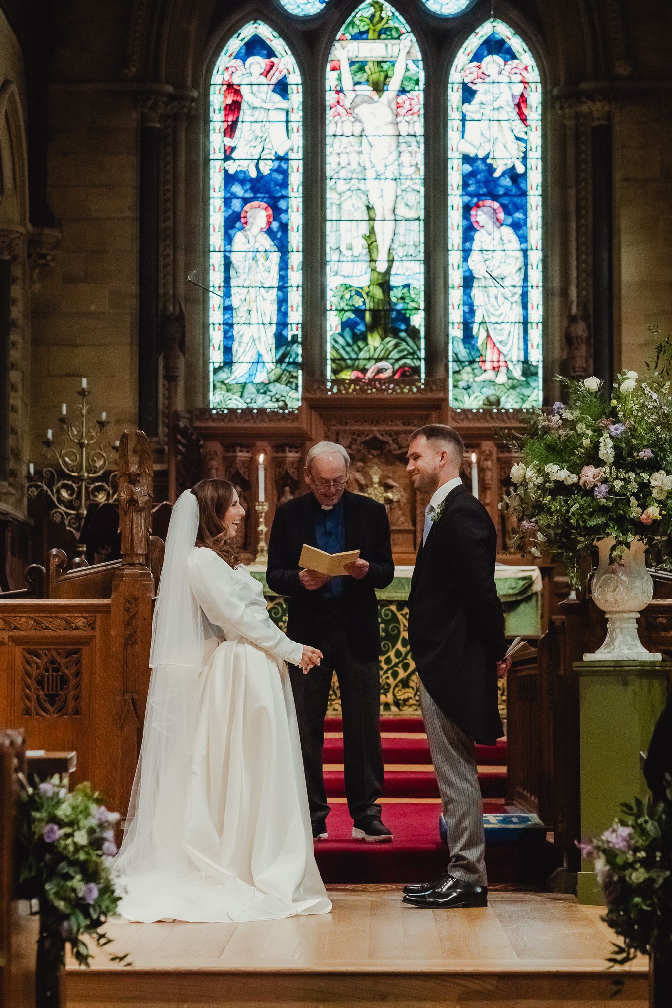 A wedding ceremony takes place in a beautiful church with stained glass windows and floral decorations.