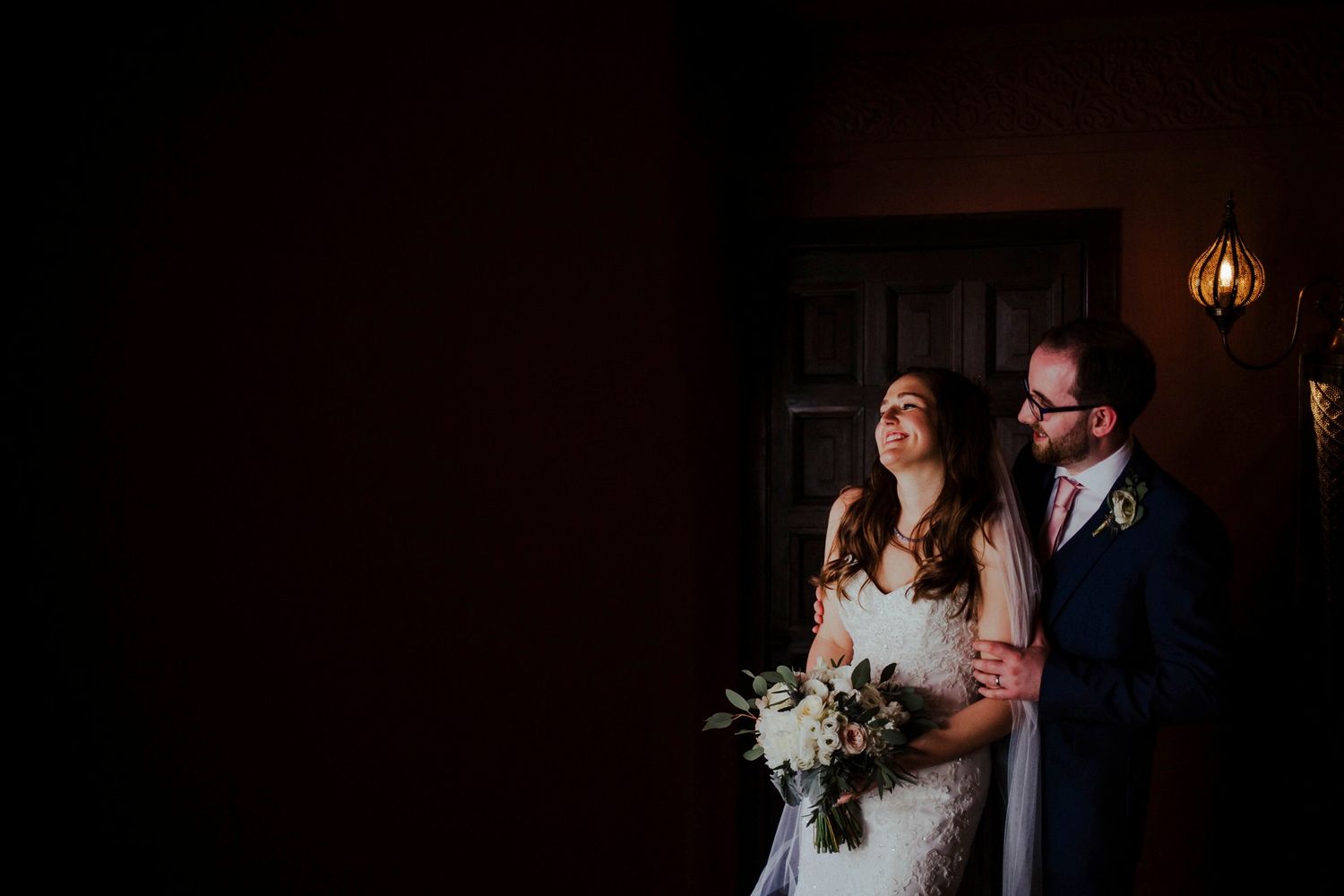 A newlywed couple stands in dramatic doorway lighting with bride holding bouquet.