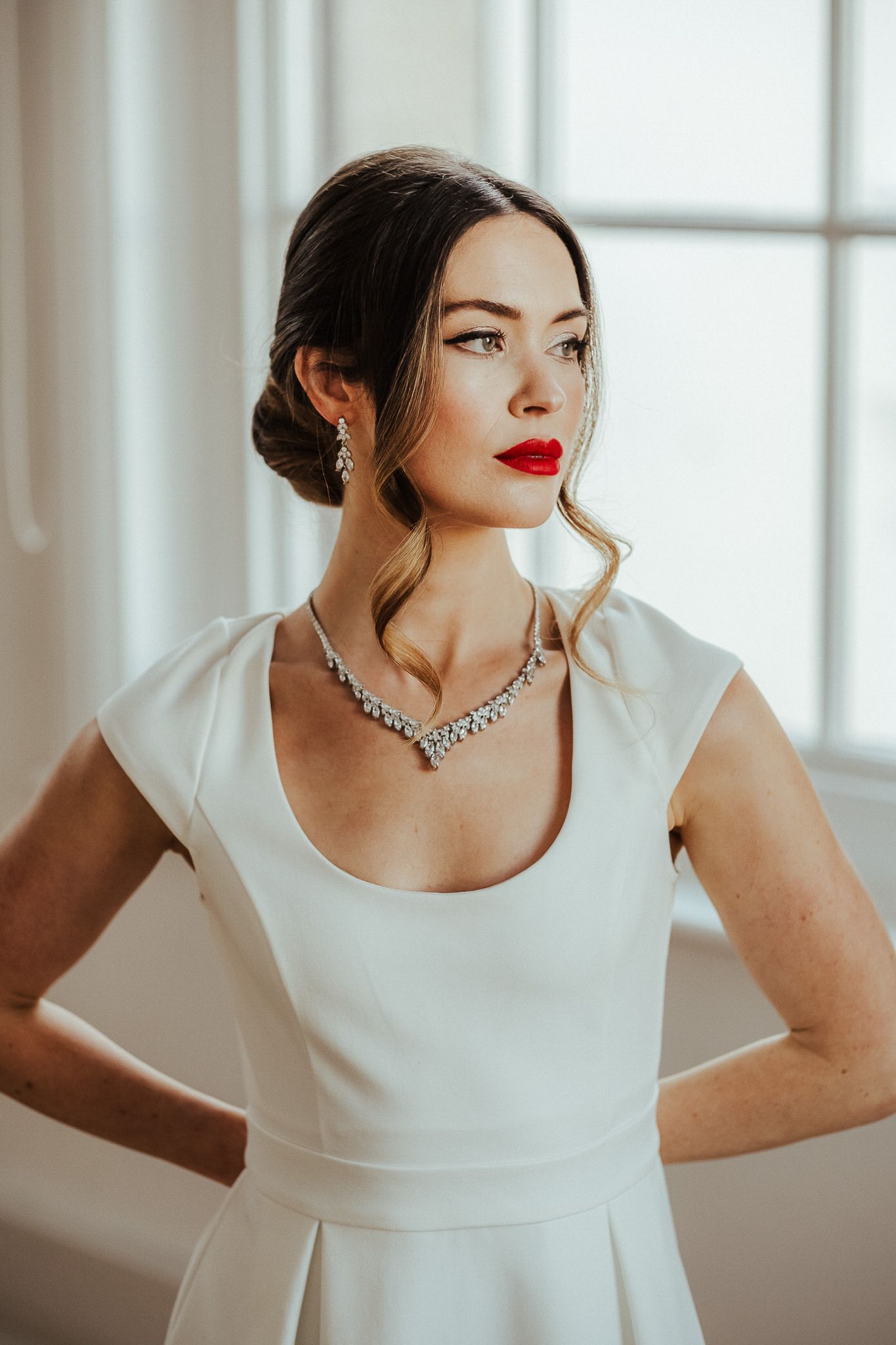 Elegant bridal portrait featuring a classic white dress, sparkling necklace, and sophisticated updo hairstyle near a window.