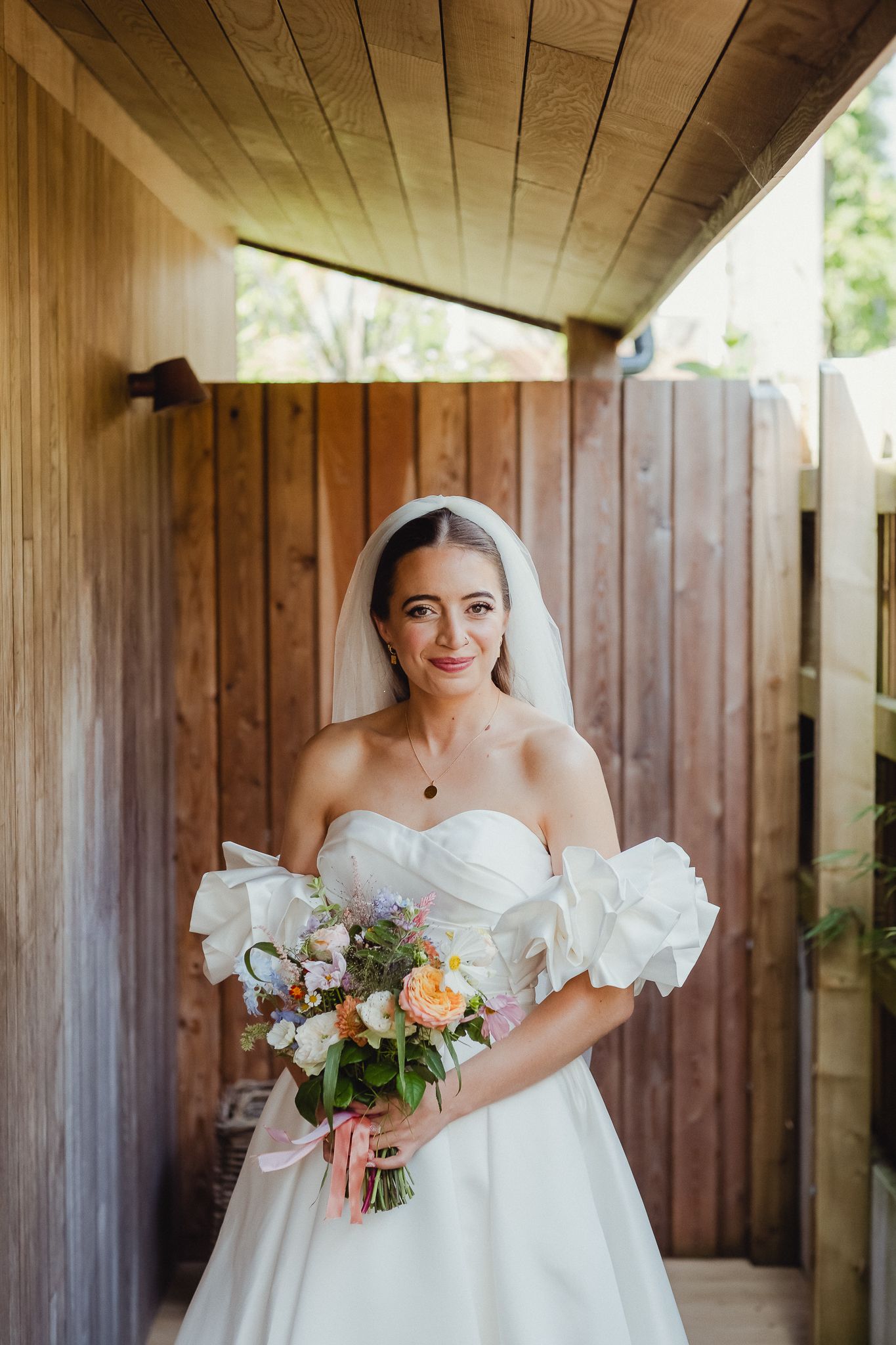 A bride in a white off-shoulder wedding dress with puff sleeves poses in a rustic wooden venue holding a bouquet.