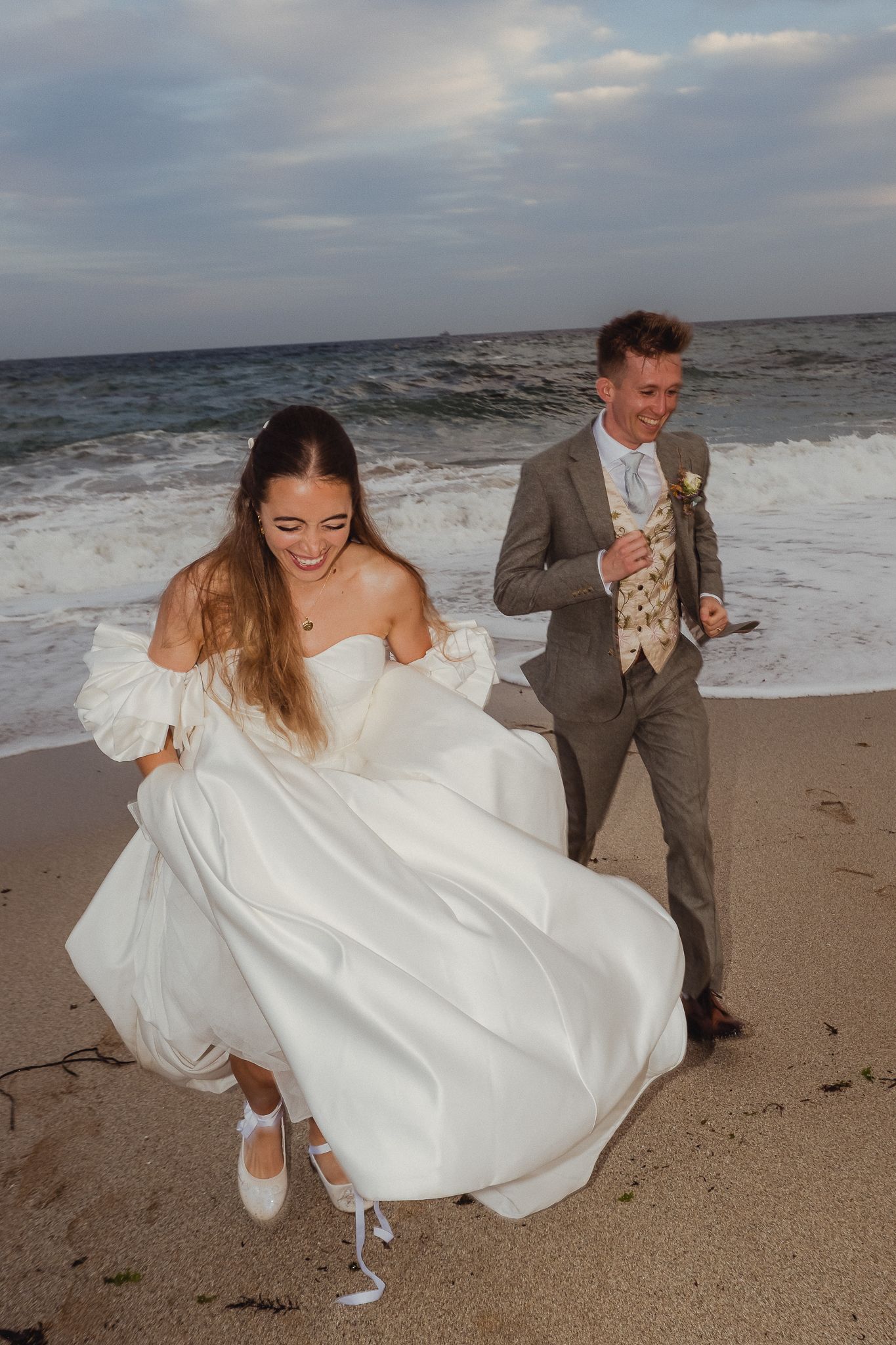 A couple in wedding attire playfully runs along a beach shore with waves crashing behind them.