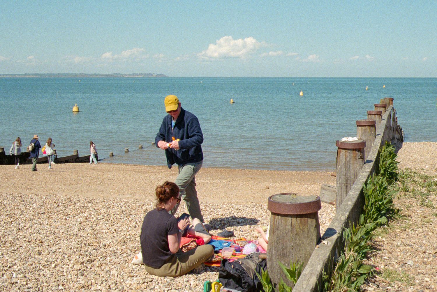 People enjoying a sunny day at a pebble beach with calm blue waters and clear skies in the background.