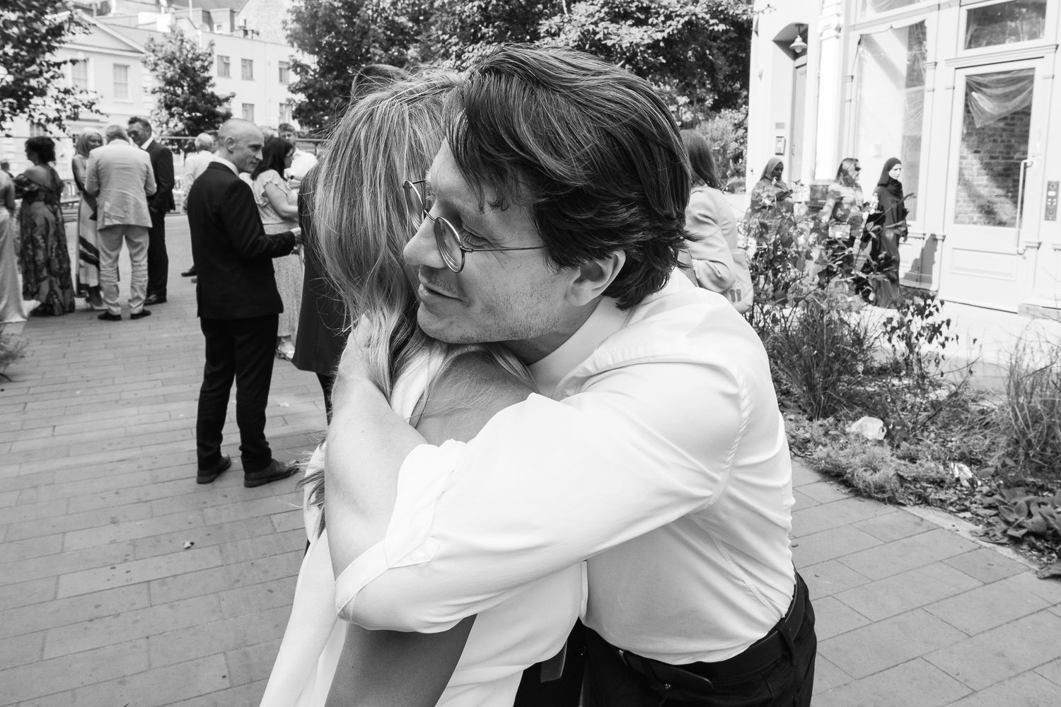 Two people share a warm embrace at an outdoor gathering while guests mingle on a brick-paved street in the background, in black and white.