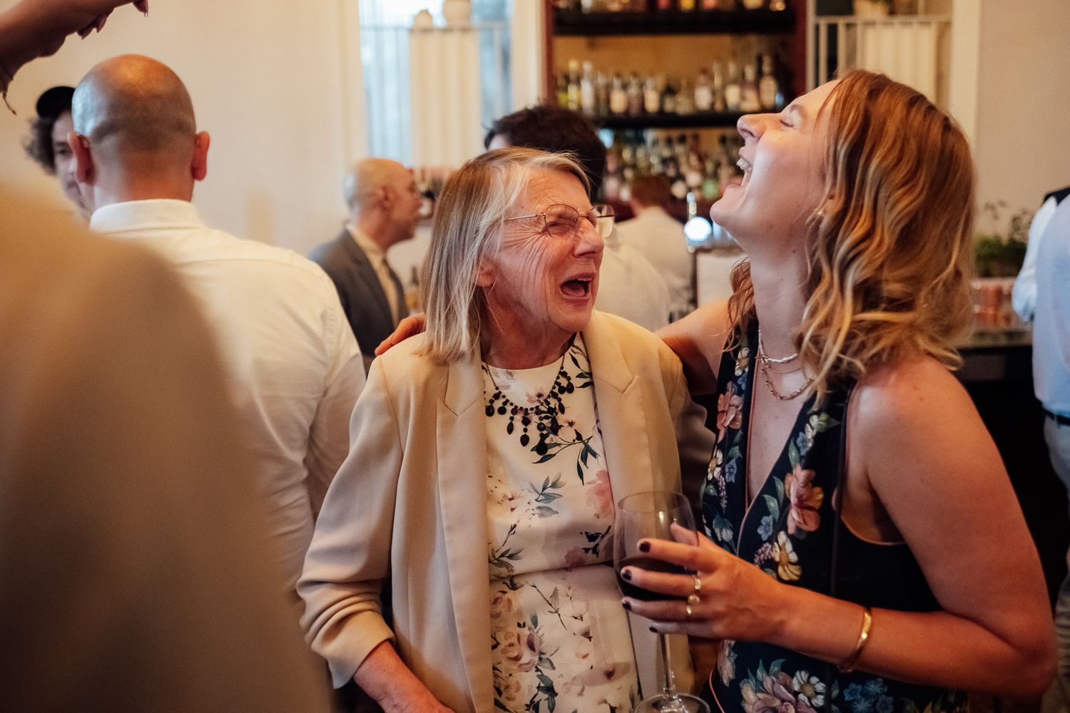 Two women laugh together at a lively indoor party, one in a floral dress holding a wine glass, with guests and a bar shelf behind them.