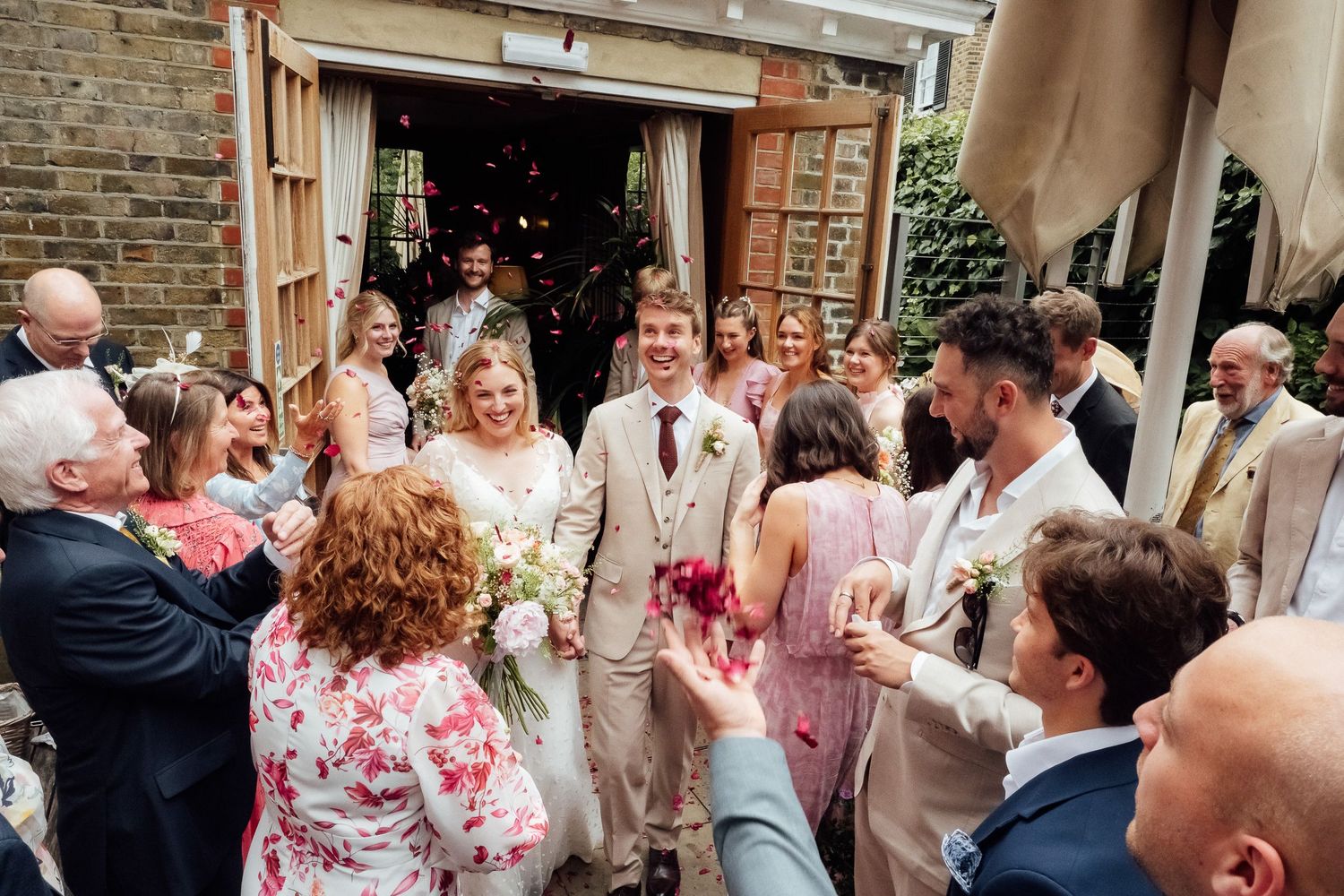 Smiling bride and groom walk through a cheering crowd of wedding guests outside a brick venue, with guests holding flowers and celebrating.