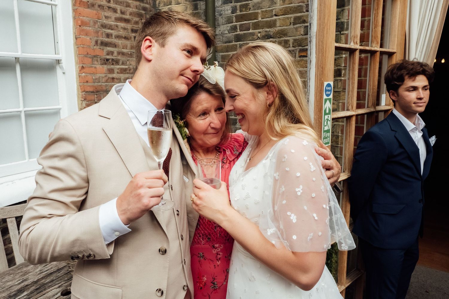 Smiling wedding guests in a warm embrace, a man in a beige suit holding champagne beside a woman in white lace, with a young man watching.