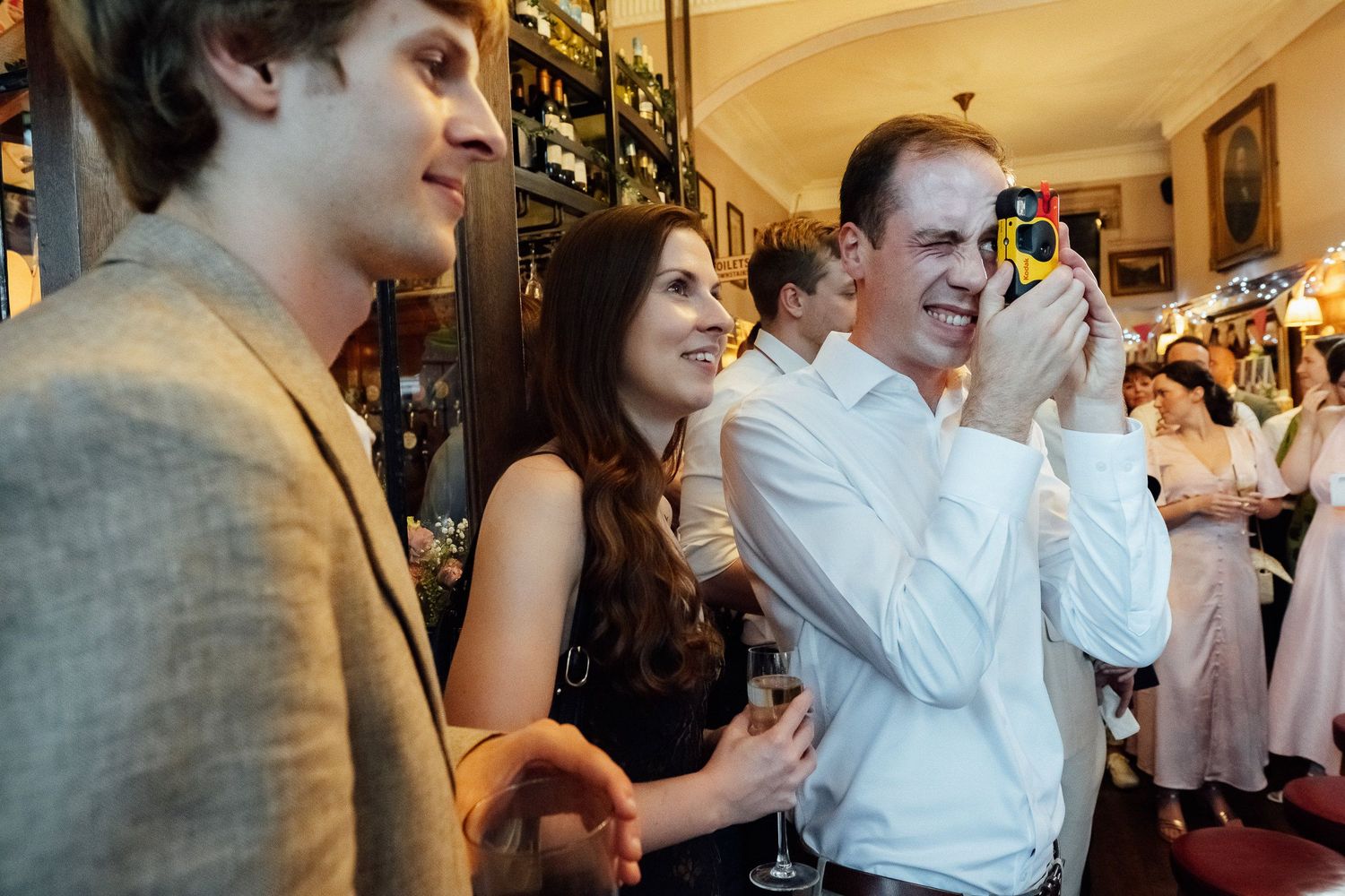 Guests mingle at an indoor party as a smiling man in a white shirt holds a camera to his eye while a woman with a drink smiles beside him.