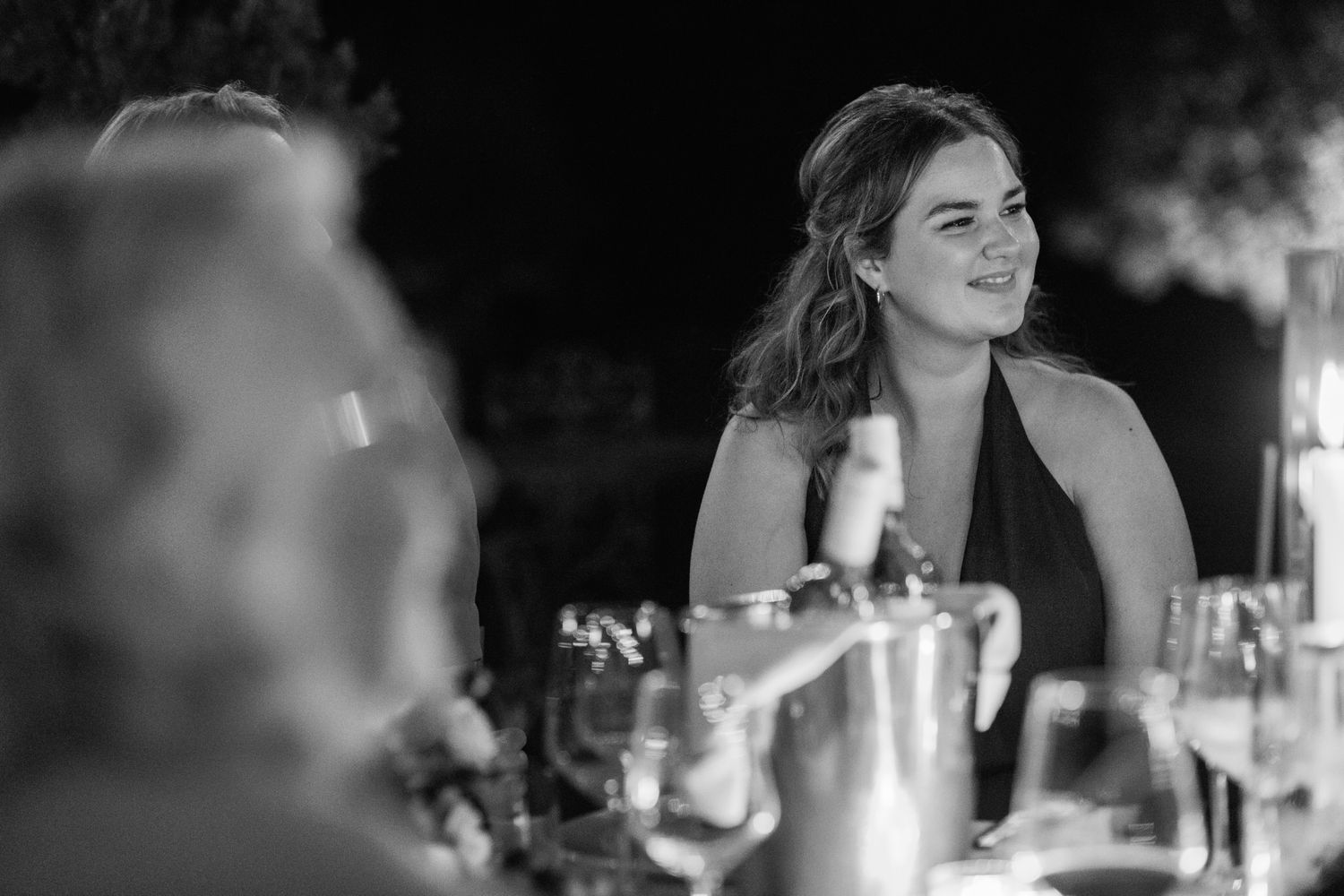 Smiling woman with wavy hair in a halter dress sits at a candlelit evening dinner table with wine glasses in black and white.