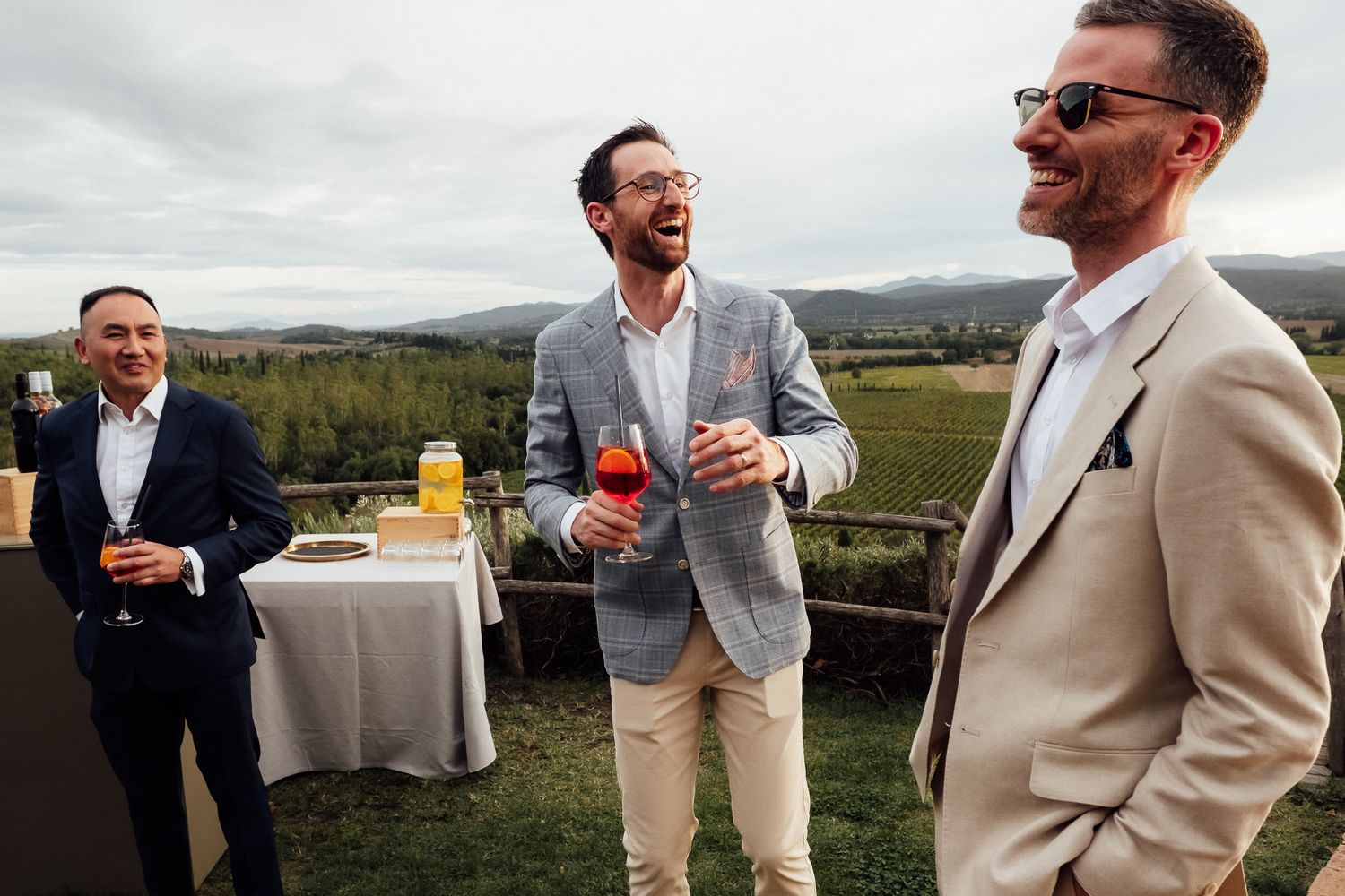 Three smiling men in suits laugh and hold drinks at an outdoor vineyard gathering with a refreshment table and rolling hills behind them.
