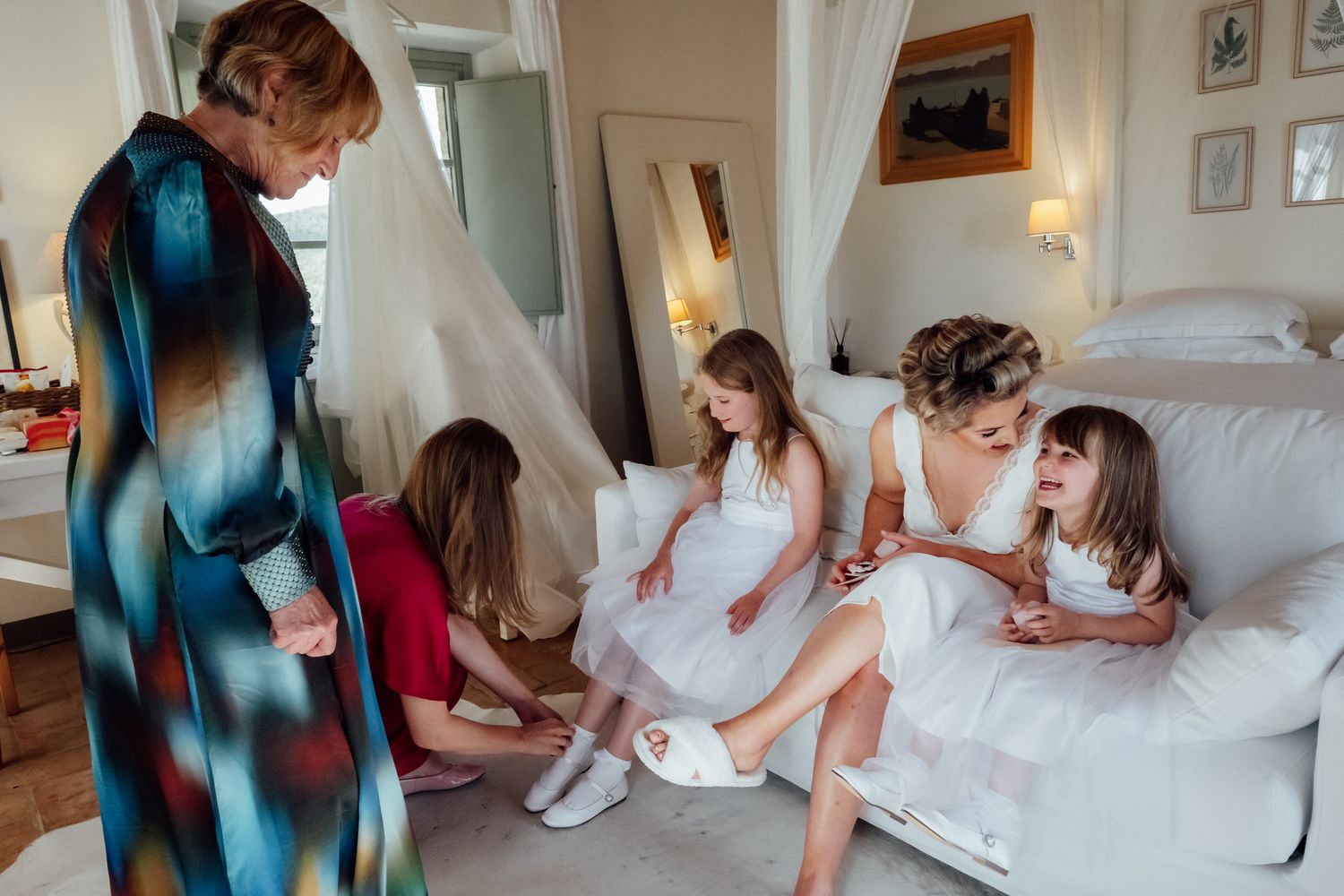 Bride in a white dress sits on a sofa with two flower girls in white dresses while two women in colorful outfits help them get ready.