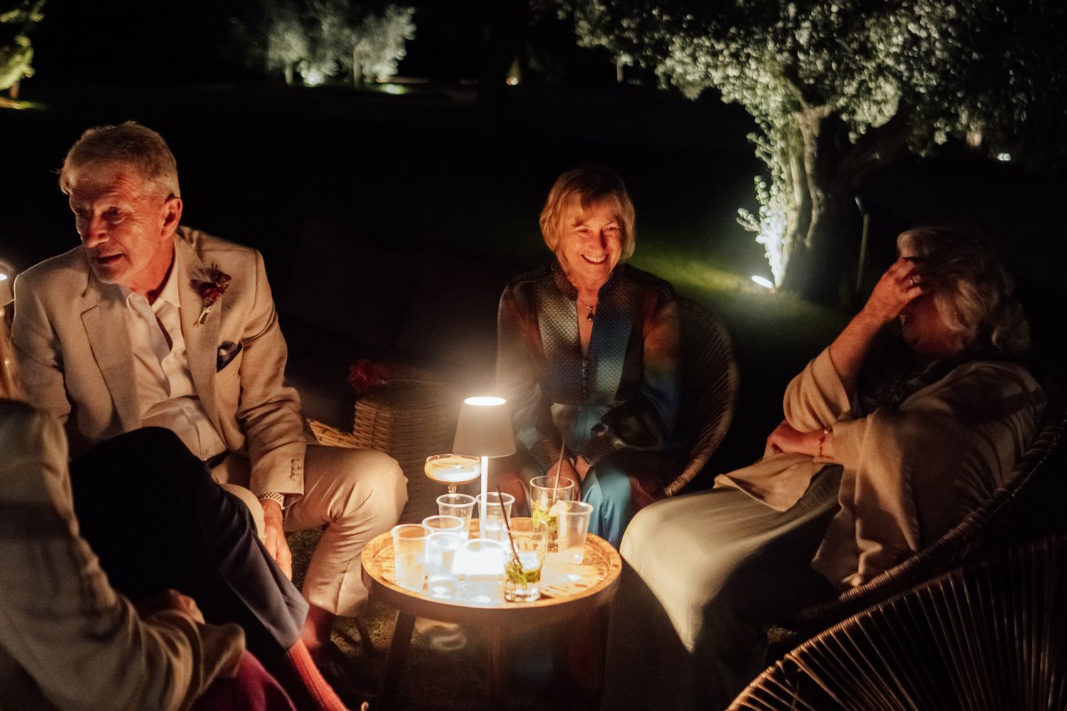 Three friends relax around a small lit table with drinks during a cozy evening outdoor gathering under softly illuminated trees.