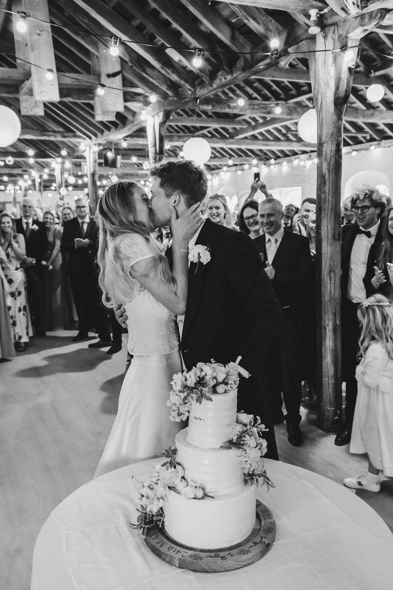 A wedding reception shows guests gathered around a rustic wooden cake display in an open-beam venue.