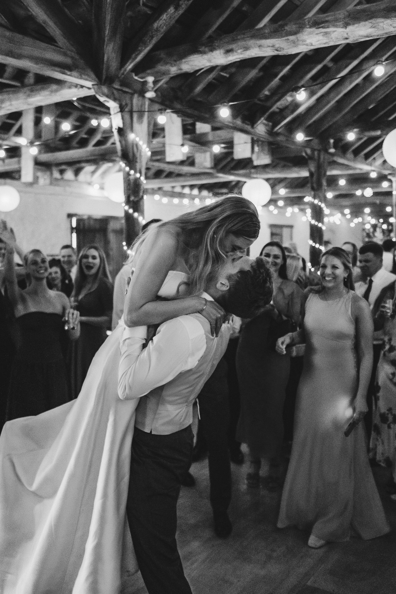 A black and white photo series showing a couple's first dance at their wedding reception under wooden beams.