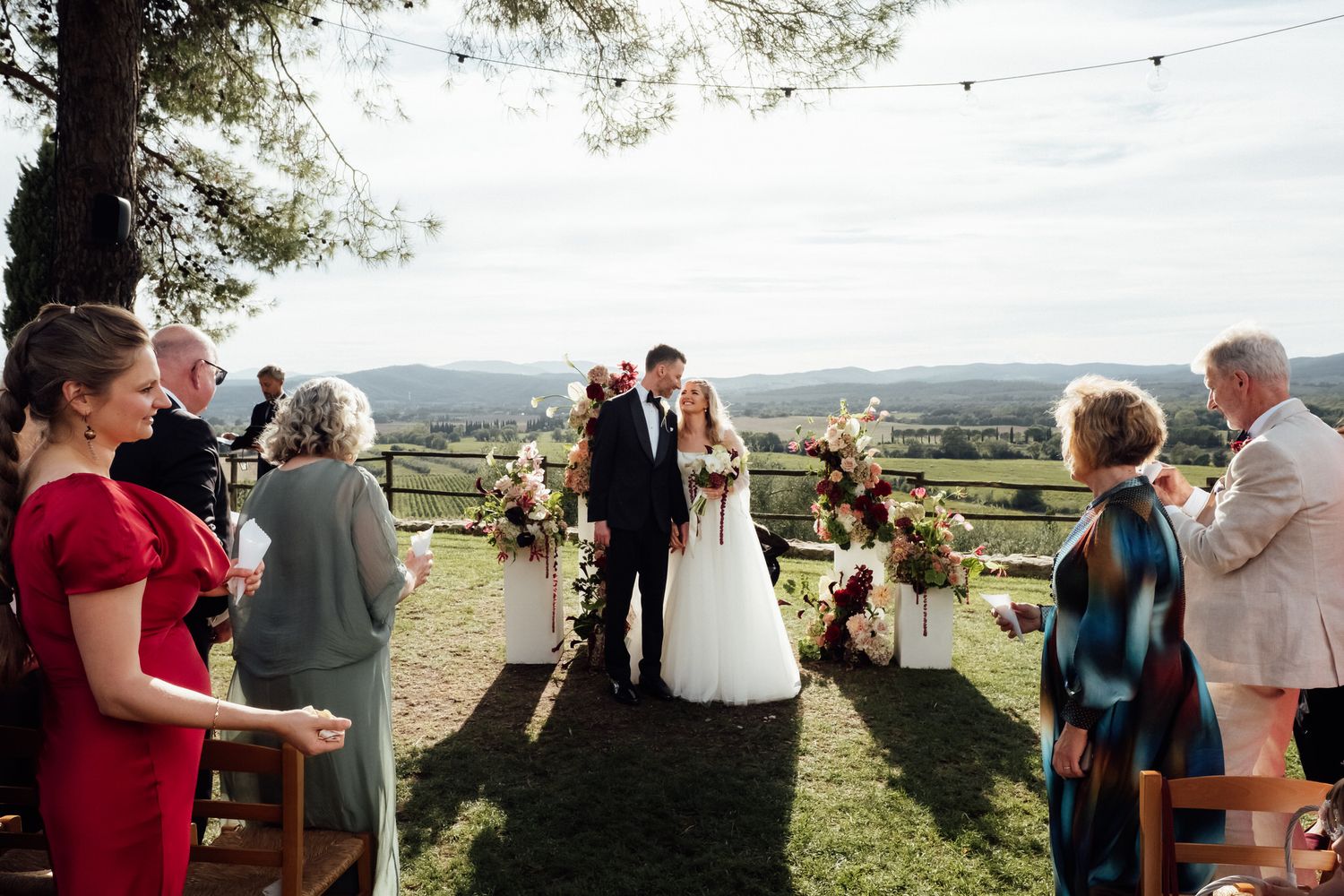 Bride and groom exchange vows at an outdoor hillside wedding ceremony surrounded by floral arrangements as guests watch.