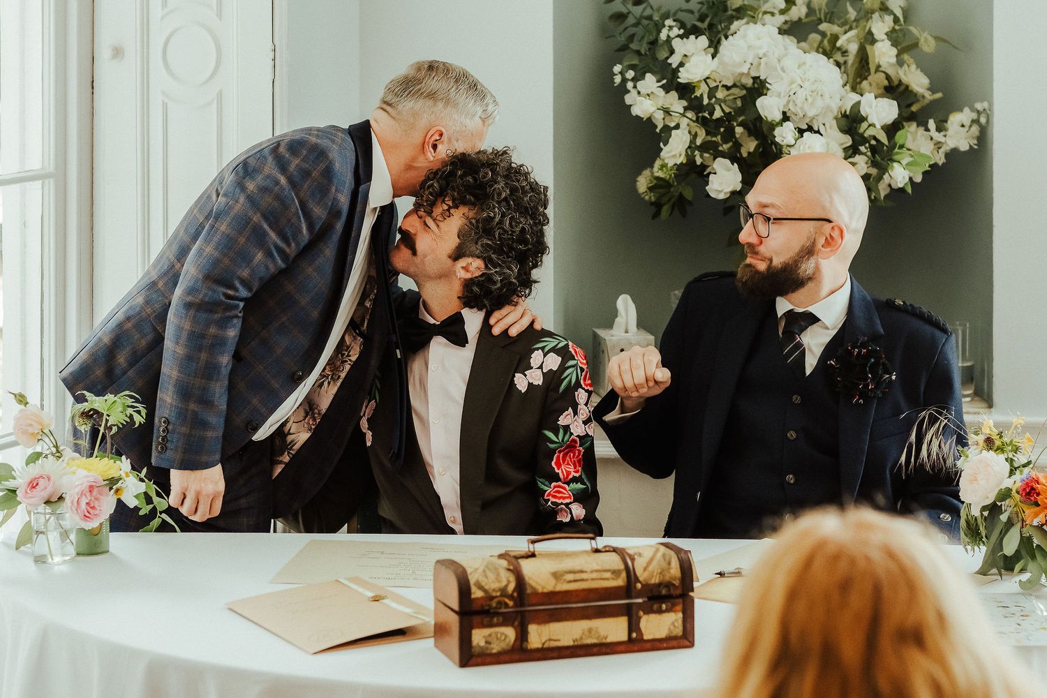 Three men in stylish suits share a joyful moment at a wedding ceremony table with floral arrangements and a wooden keepsake box.
