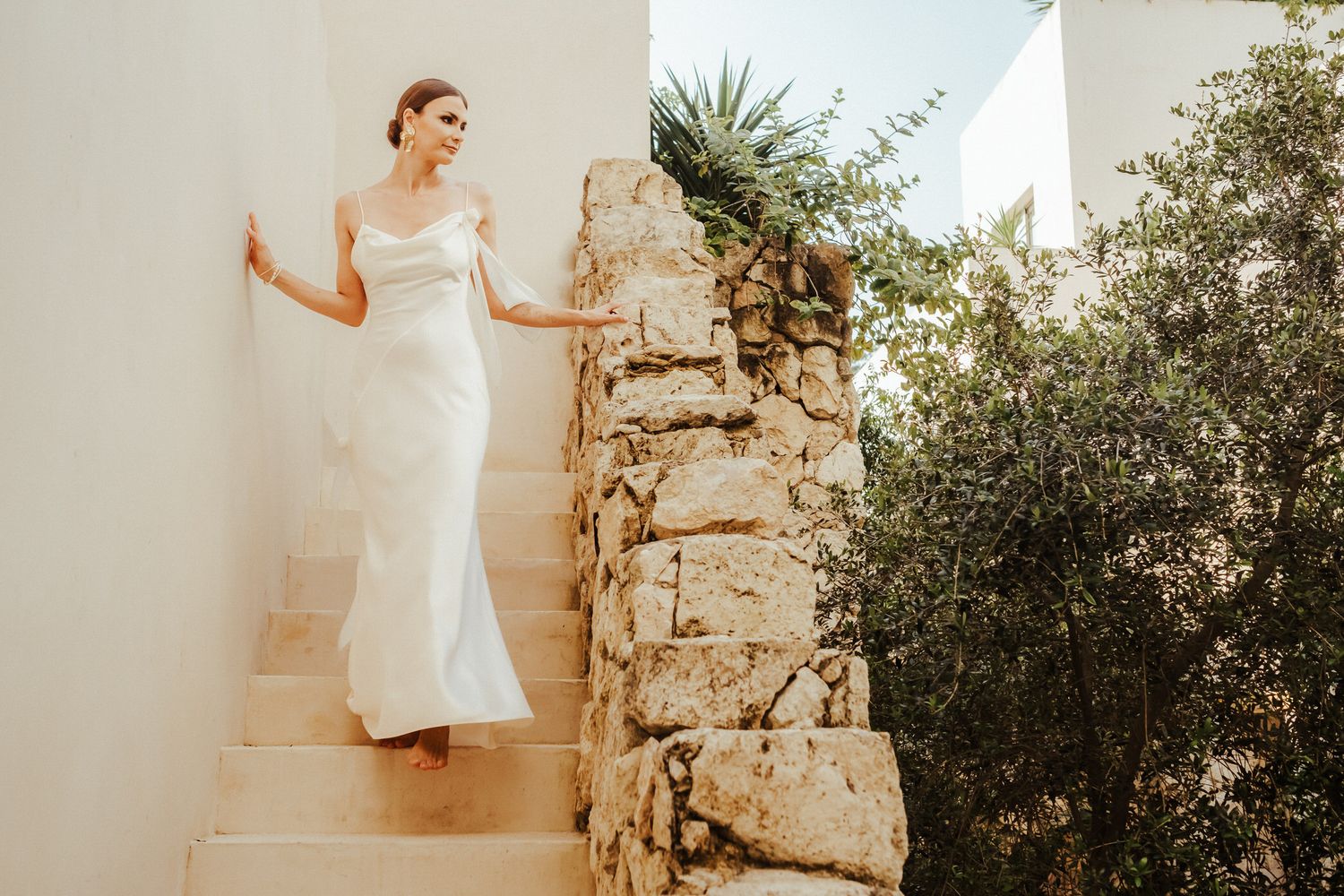 A bride in a white wedding dress poses elegantly against a rustic stone wall in a garden setting.