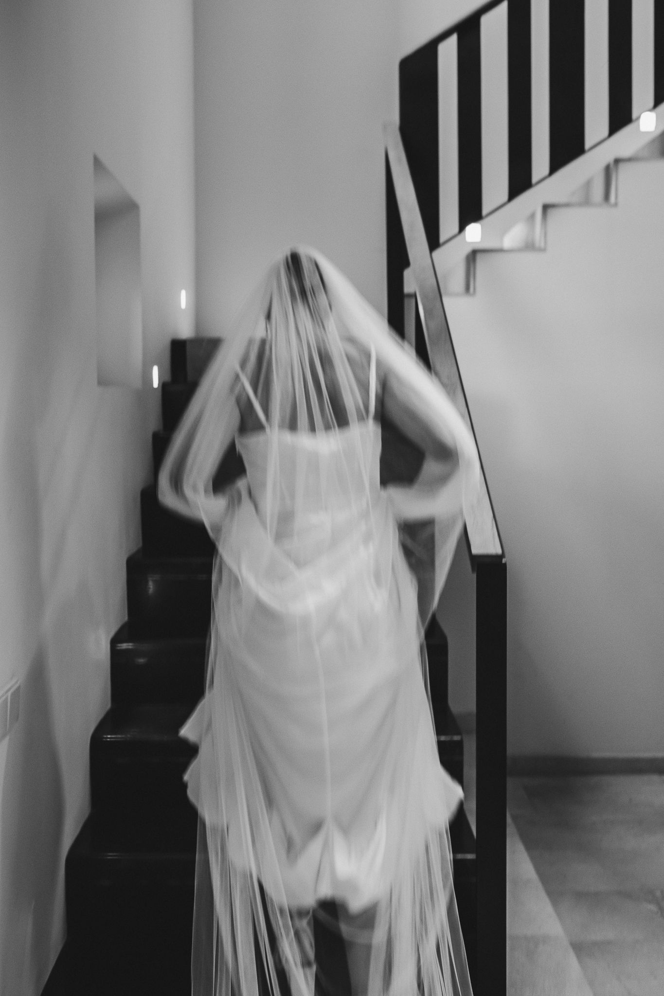 A bride in a flowing white dress and long veil ascends a modern staircase in black and white photography.