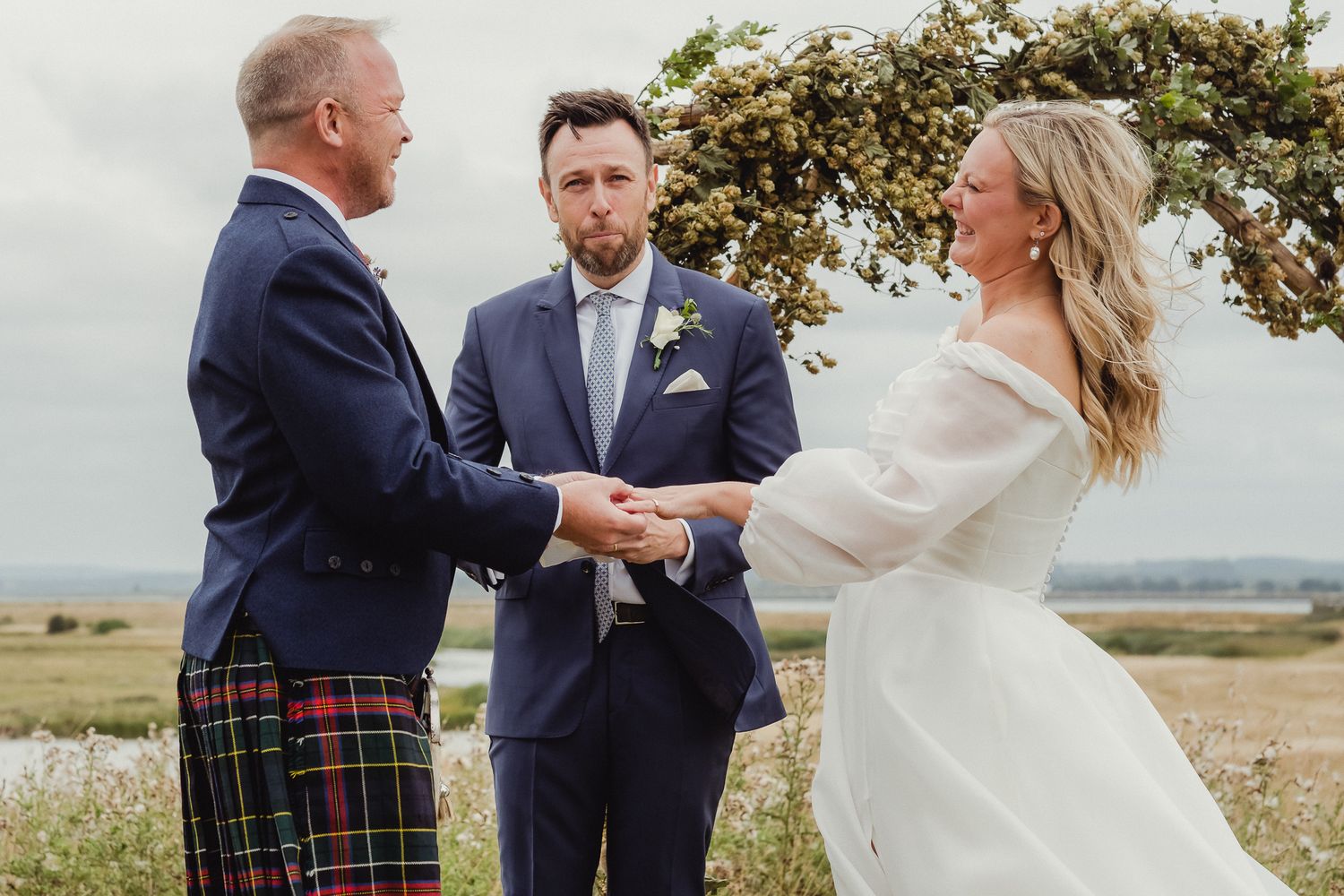 A couple shares an intimate moment in a field while wearing wedding attire including a white dress and plaid kilt.