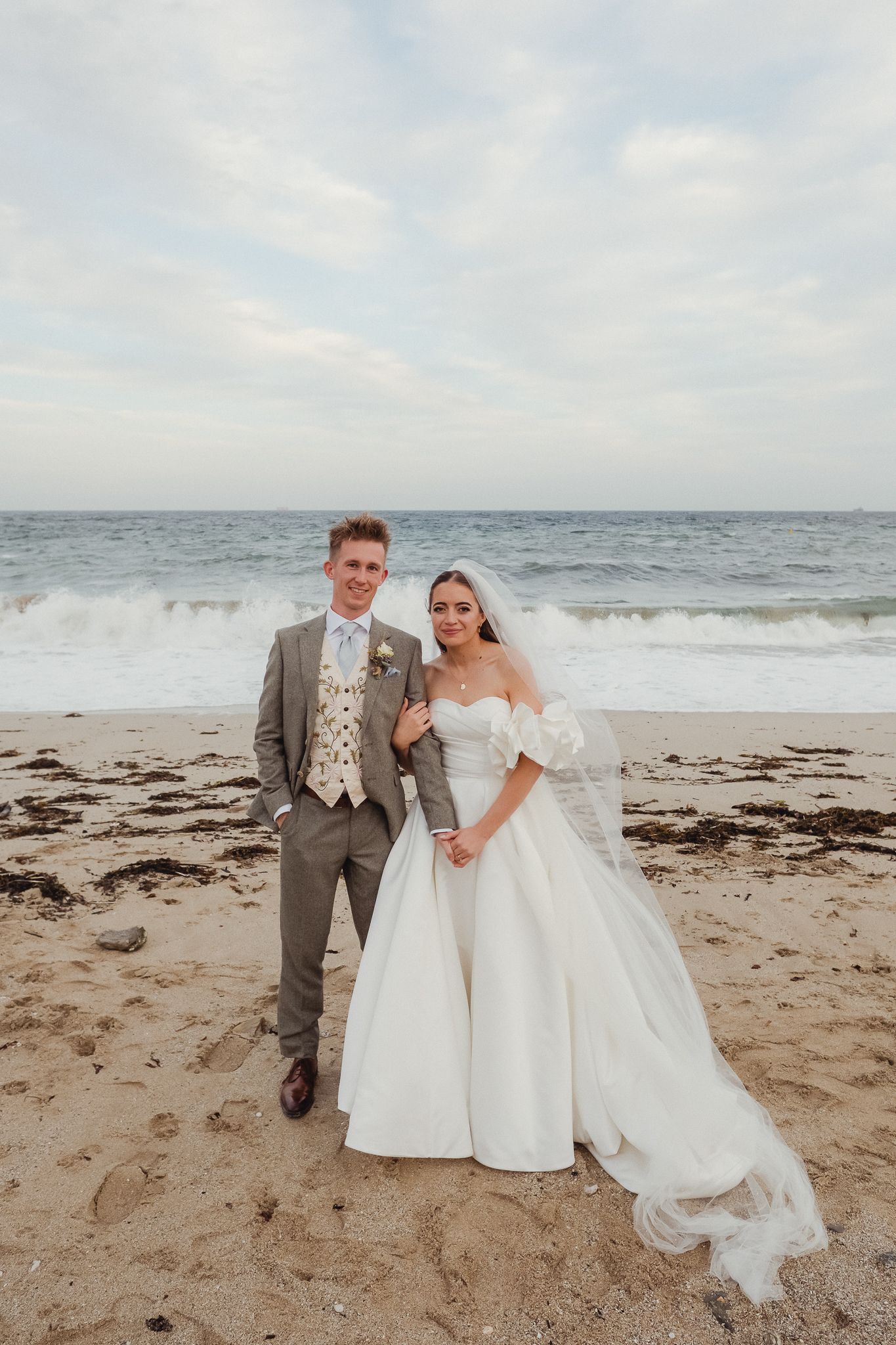 A couple poses for wedding photos on a sandy beach with ocean waves and cloudy skies in the background.