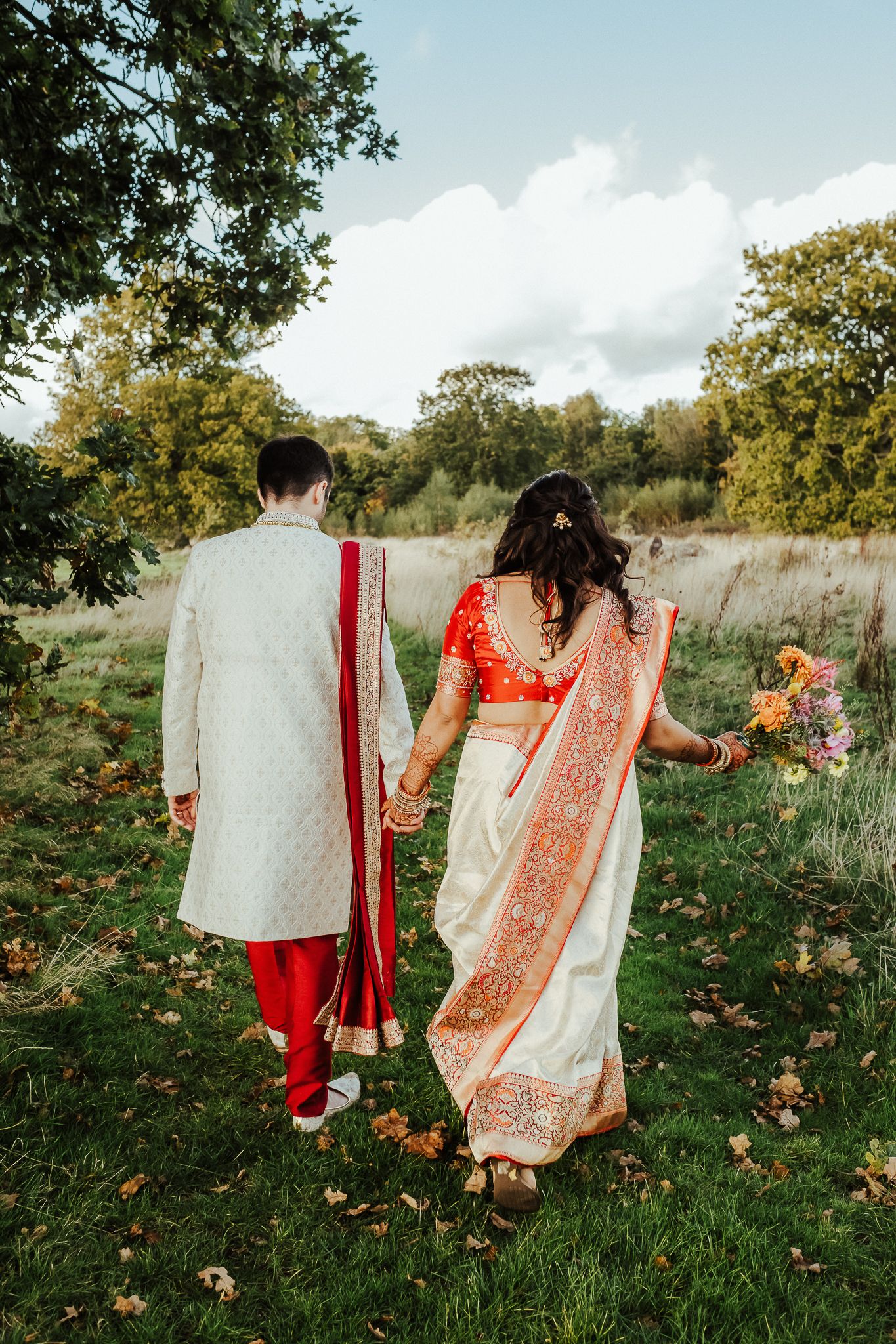 Couple in traditional Indian wedding attire walking hand in hand through an autumn field.