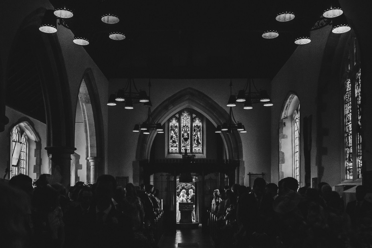 Dark church interior during wedding ceremony with dramatic lighting and arched stained glass windows.