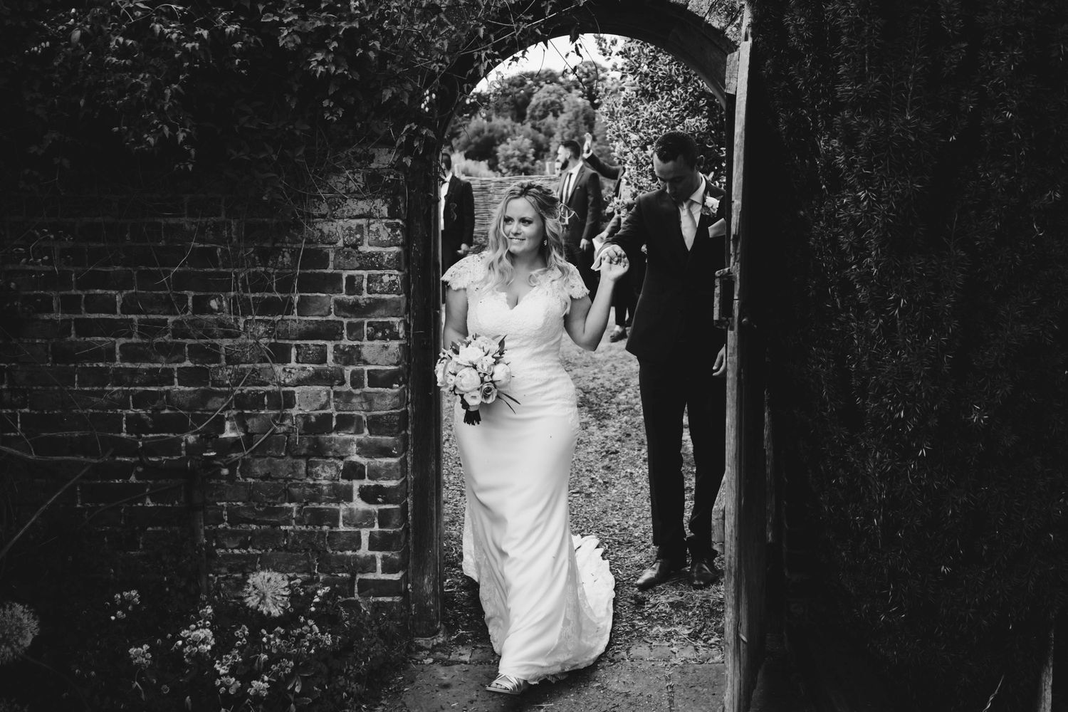 A bride emerges through a brick archway carrying a bouquet in black and white.