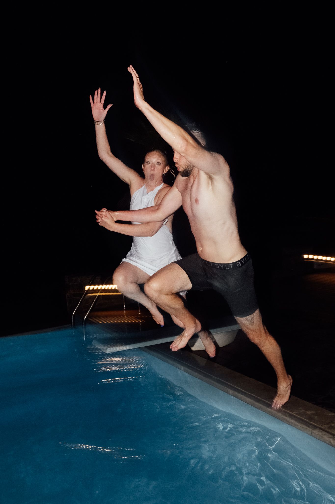 Couple jumping into a swimming pool at night, arms raised in excitement, wearing casual clothes.