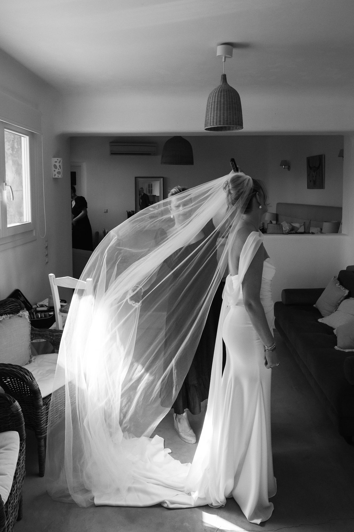 Black and white photo of a bride in a flowing gown with veil billowing gracefully inside a sunlit home.