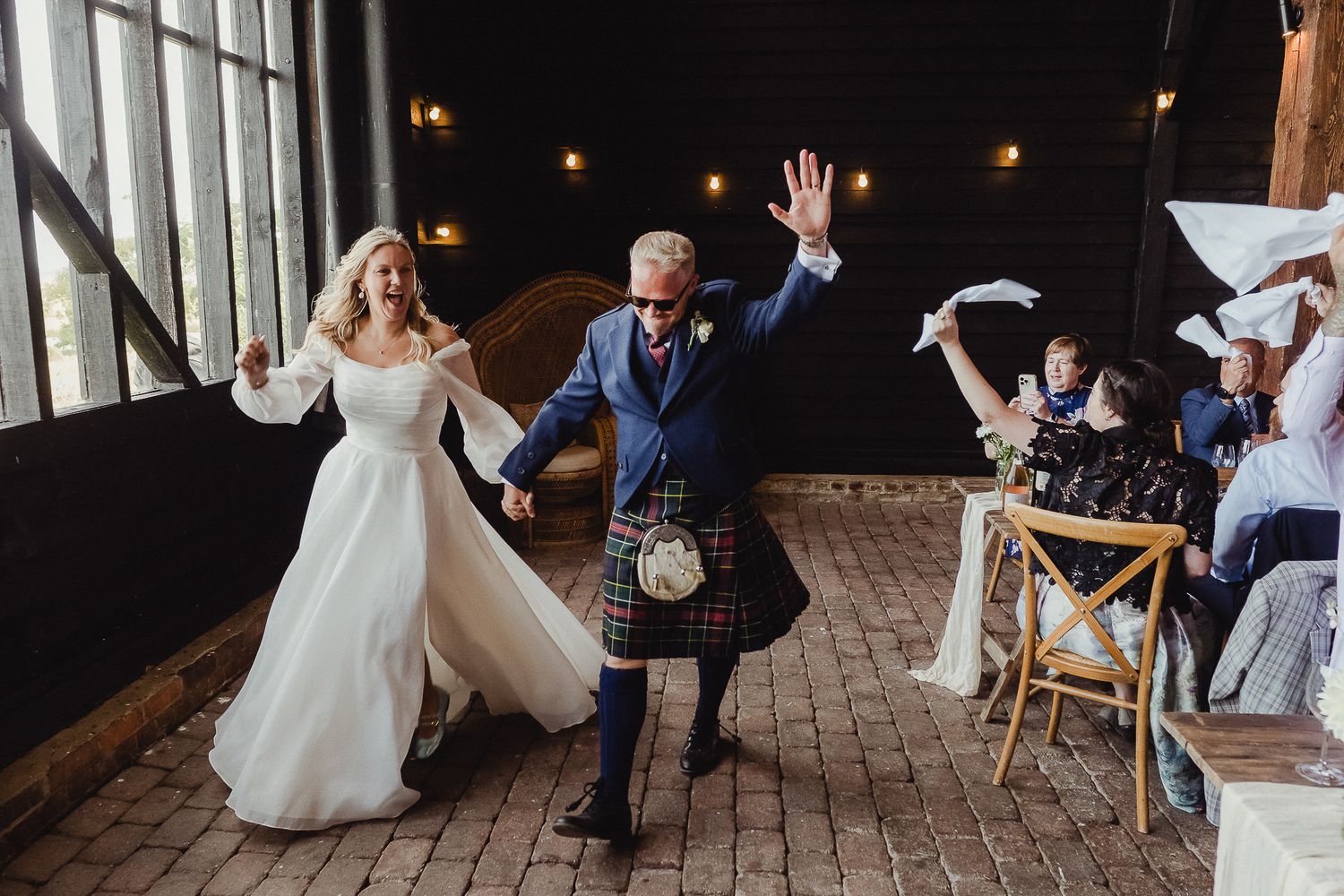 Wedding guests dance and celebrate on a rustic wooden deck with string lights overhead during an evening reception.