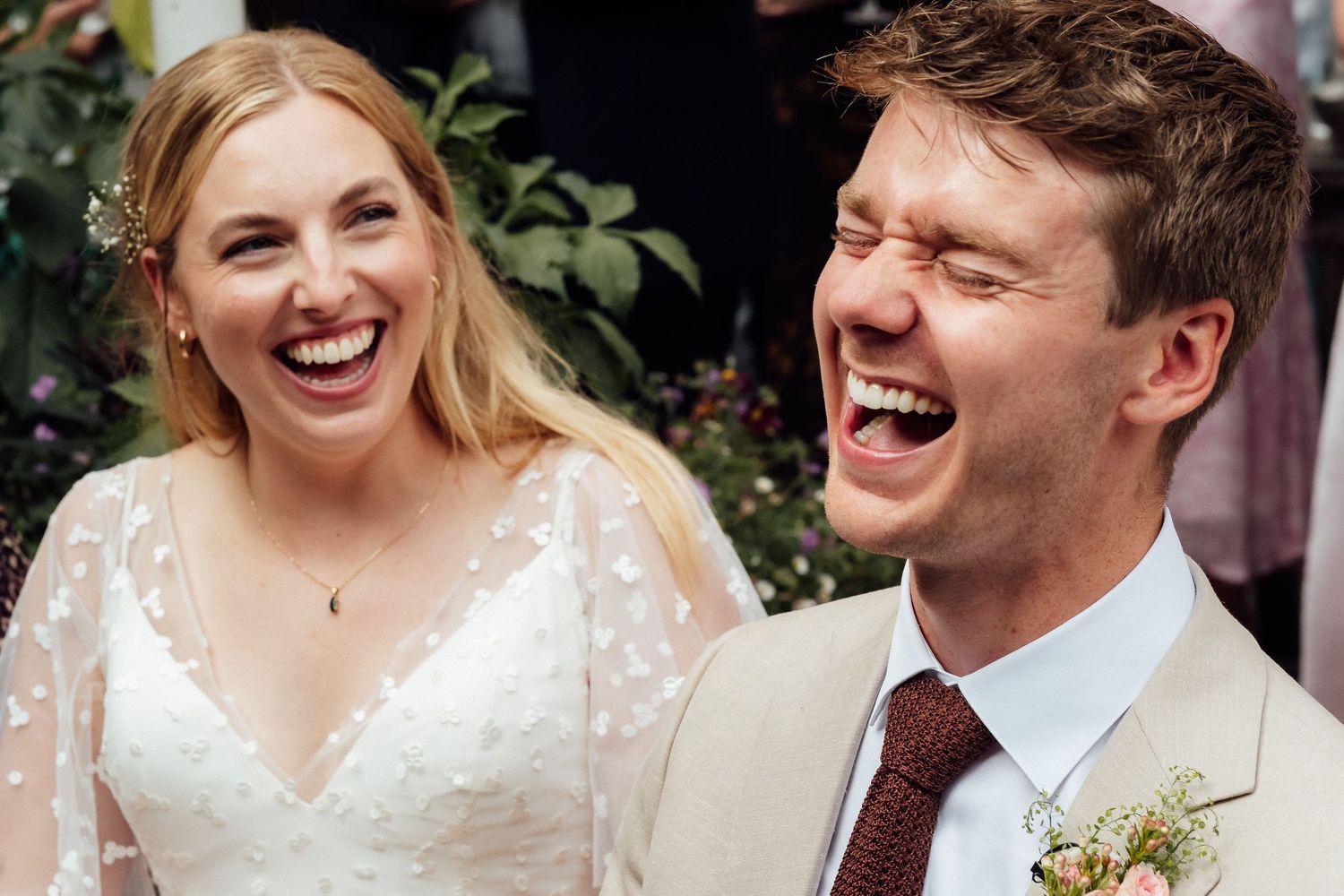 Laughing bride in a white lace wedding dress and groom in a beige suit with a burgundy tie and boutonniere at an outdoor wedding.