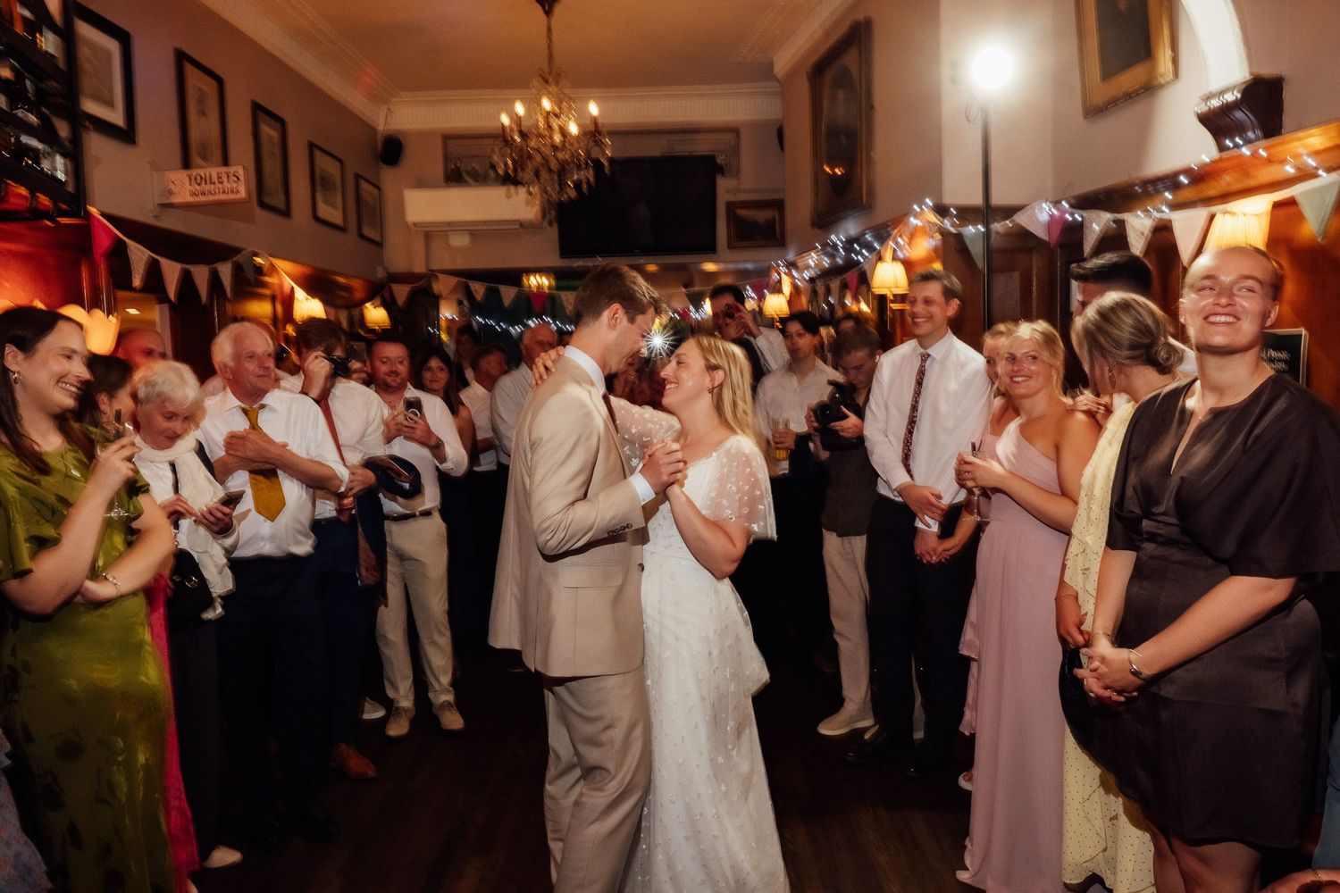 Bride in a white gown and groom in a gray suit share their first dance as smiling wedding guests watch in a warmly lit, festive venue.