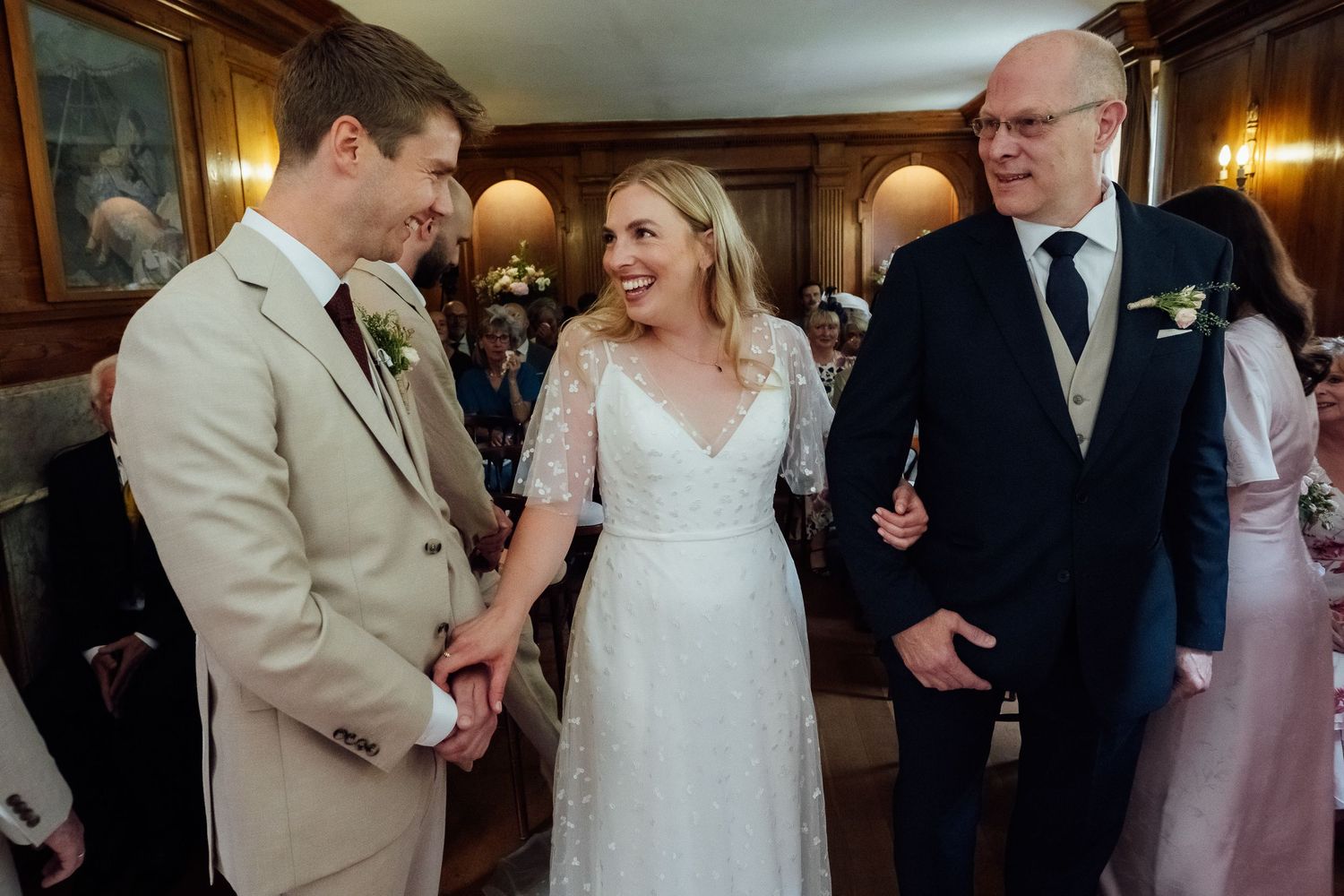 Smiling bride in a lace wedding dress holds hands with a groom in a tan suit as an older man in a navy suit looks on in an elegant venue.