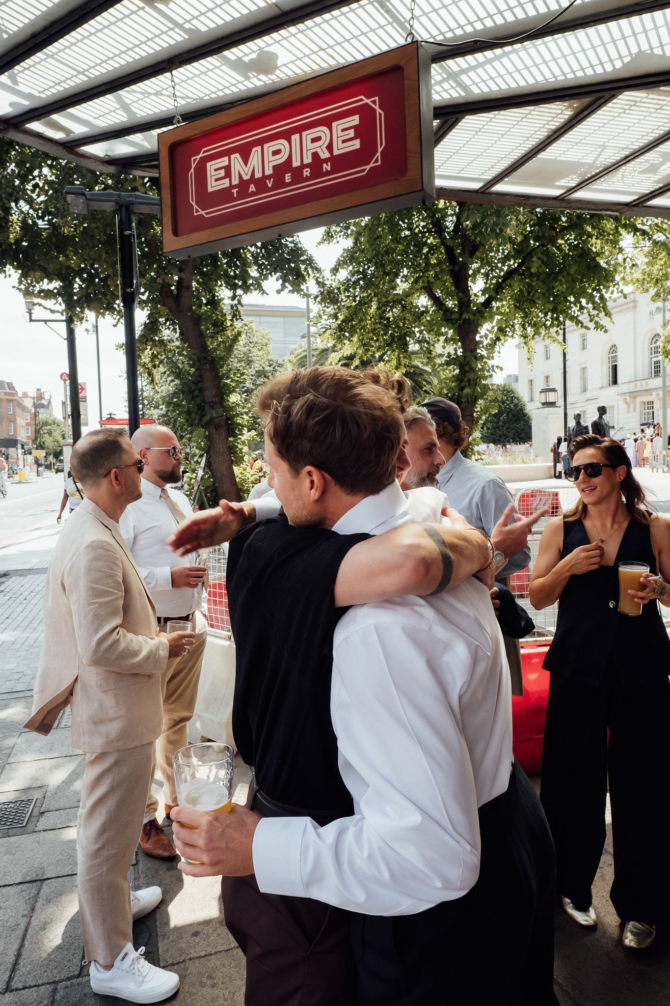 Two men hug outside Empire Tavern while friends hold drinks and chat on a sunny, tree-lined city street.