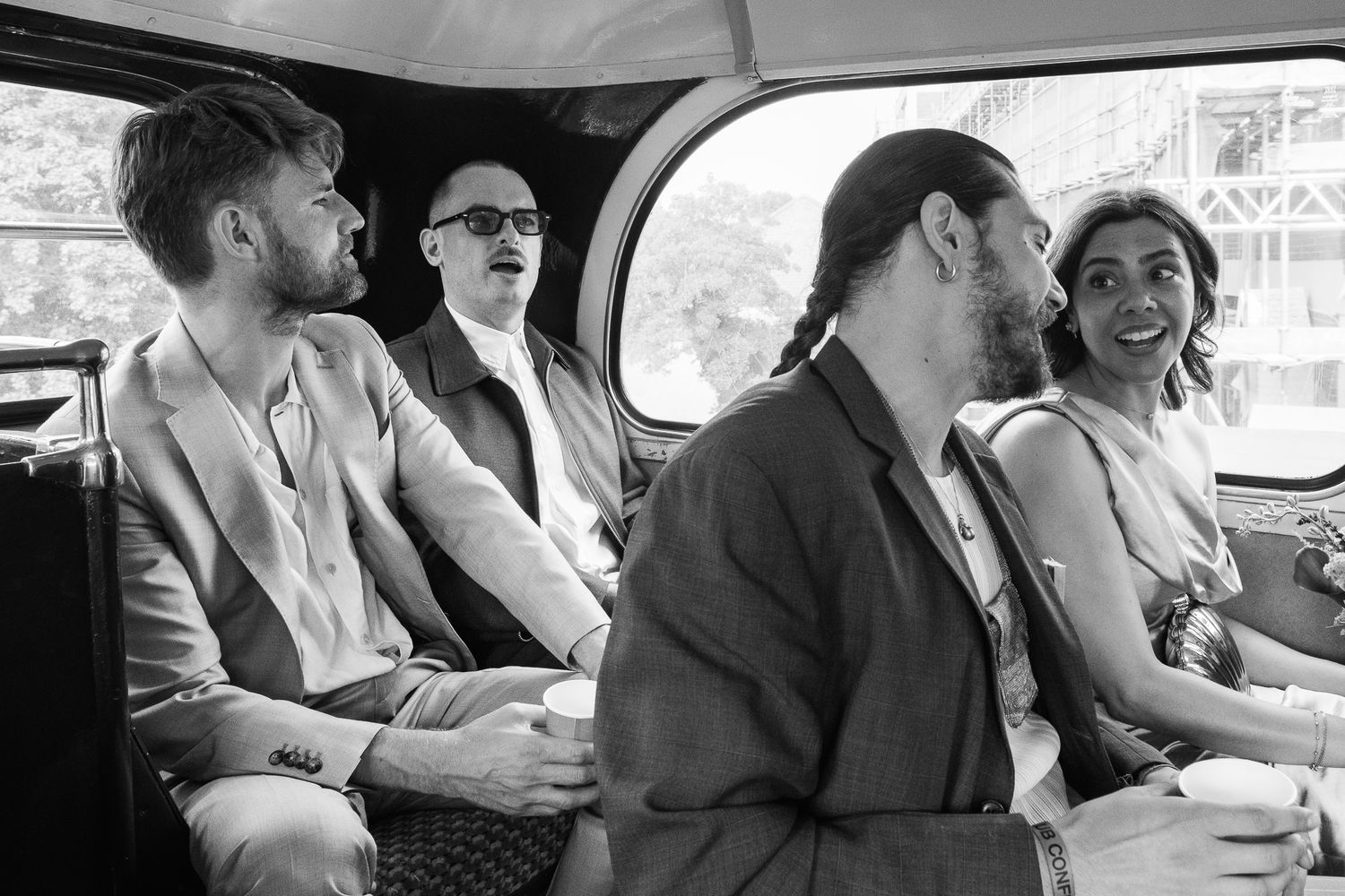 Four stylish people in blazers sit and chat on a vintage bus in a black and white photo, one holding a paper cup.