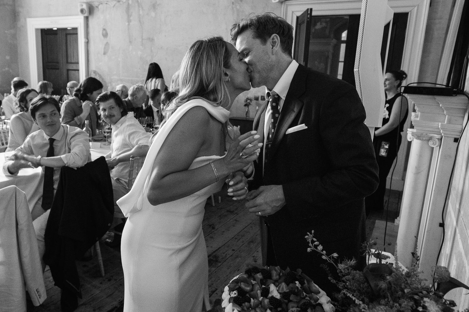 Bride in a white gown and groom in a dark suit share a kiss at a wedding reception while seated guests look on in black and white.