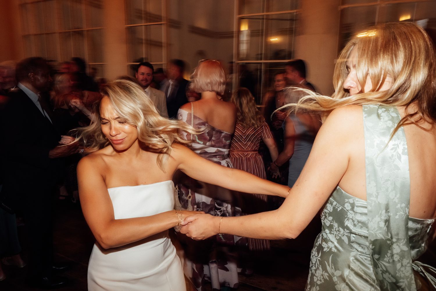 Two women with flowing hair dance and hold hands at a lively indoor party while guests mingle in the warmly lit background.