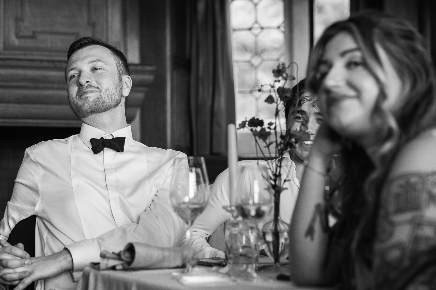 Black and white photo of guests smiling and laughing at a wedding reception dinner table with flowers.