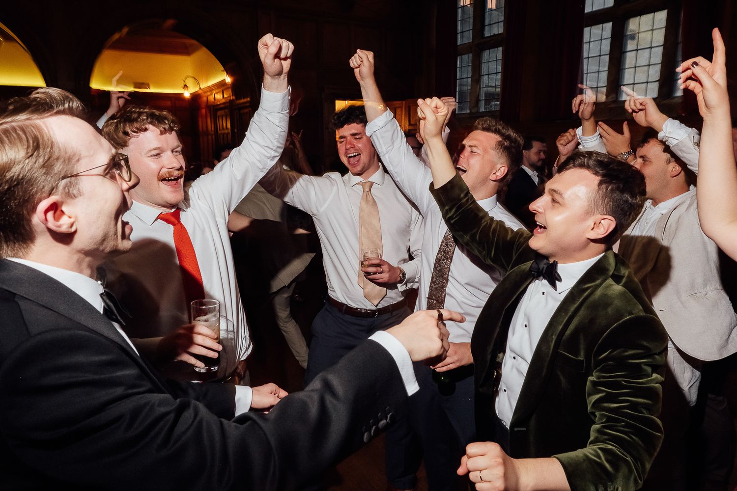 Excited groomsmen celebrating at a wedding reception, raising fists and cheering in a grand venue.