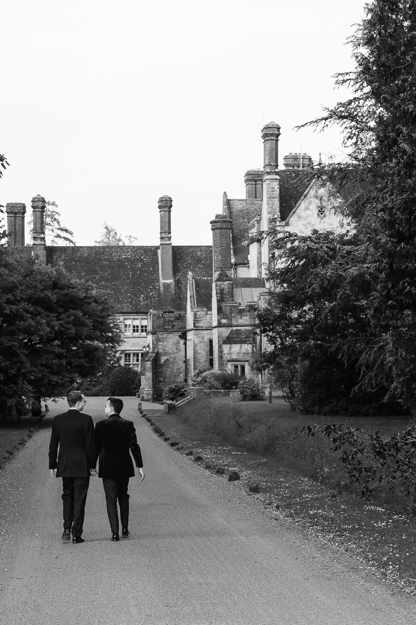 Two men in suits walk along a path toward a grand historic manor house, surrounded by trees, in black and white.