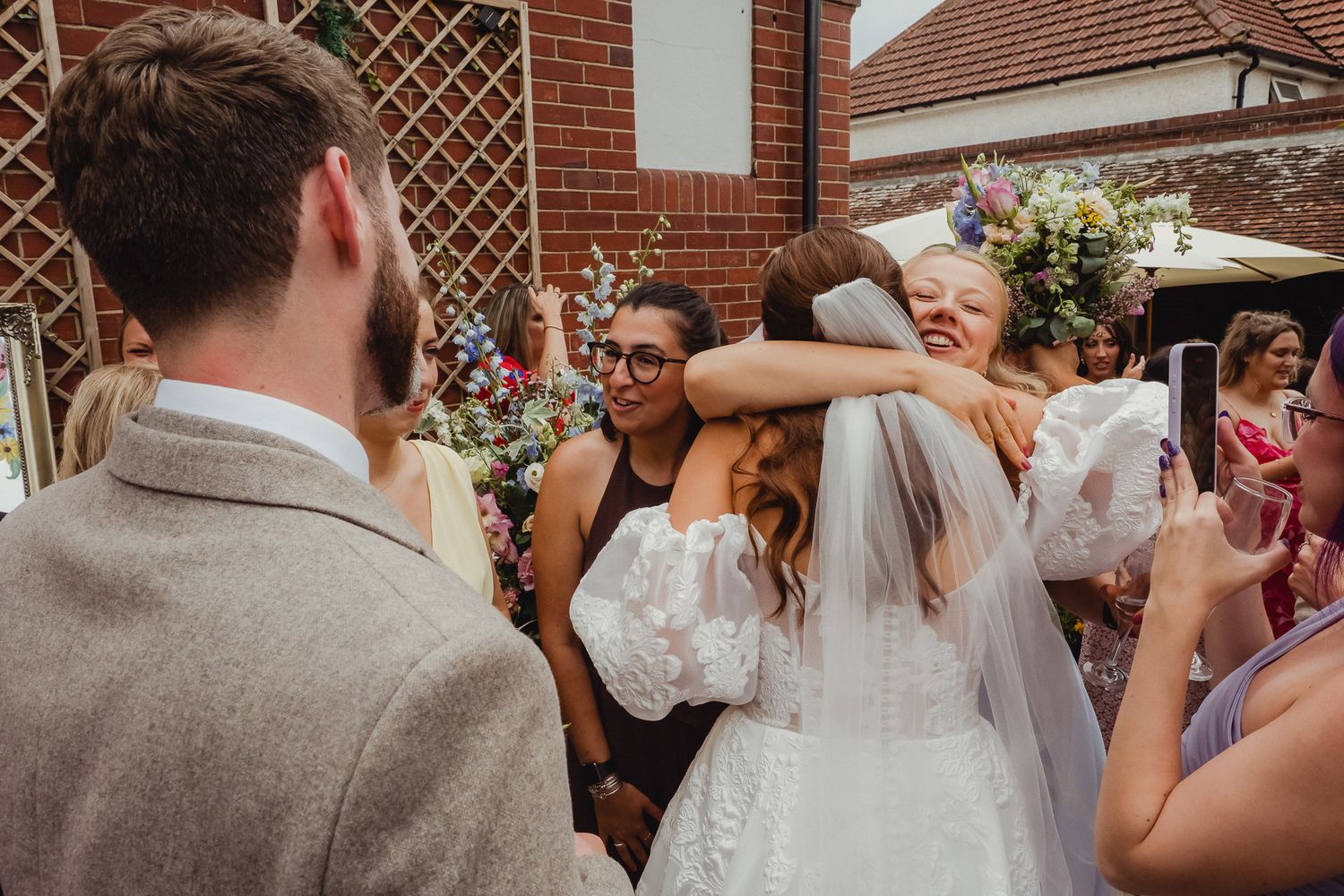 Wedding guests celebrate with hugs and smiles outside a brick building with floral decorations.