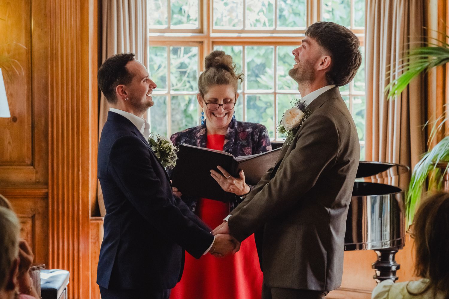 Group of people in formal attire laughing and socializing at an elegant wood-paneled room with large windows.