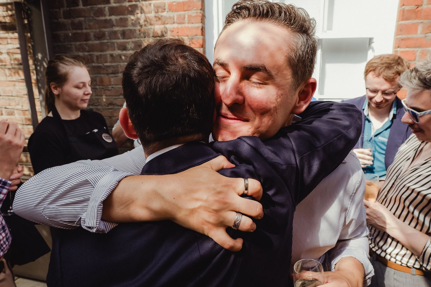 Wedding guests share emotional embraces and hugs during a celebration at a brick-walled venue.