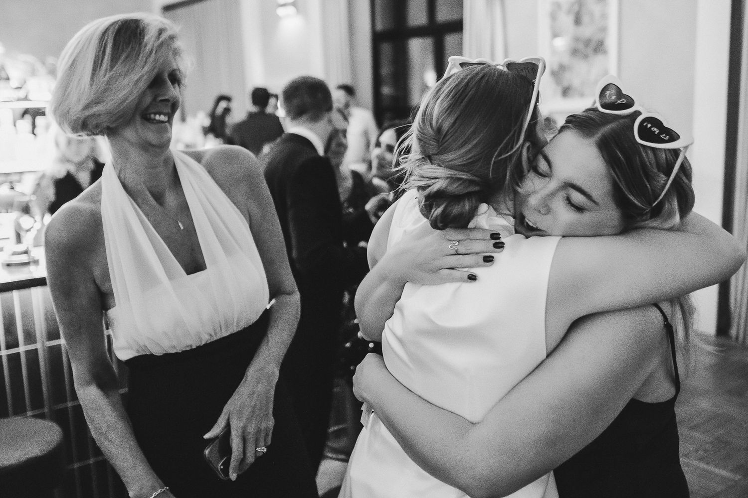 A black and white candid photo of people embracing at an indoor celebration while another person looks on.
