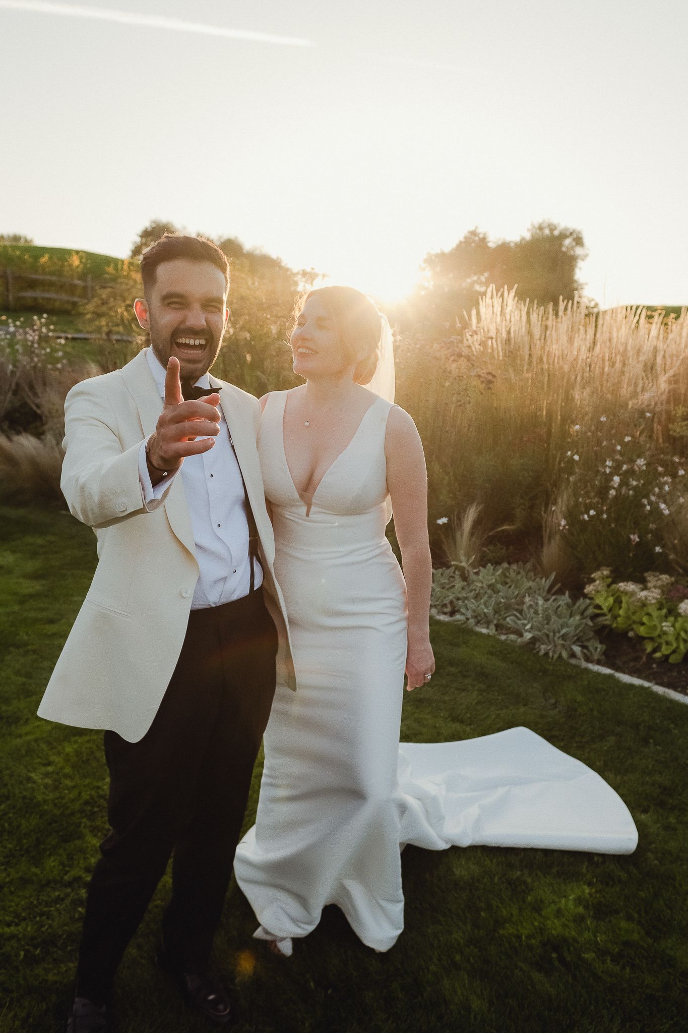 A couple in white wedding attire shares a joyful moment in a sunlit garden during golden hour.