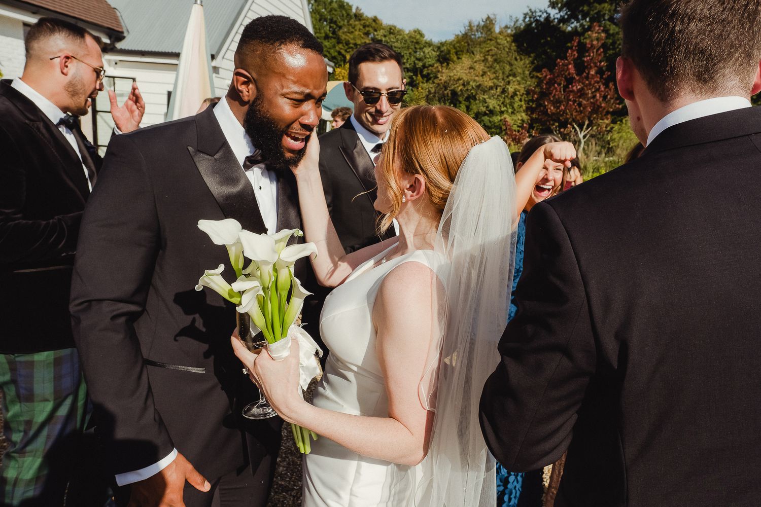 Joyful wedding celebration outdoors with bride holding white calla lilies while surrounded by guests in formal attire.